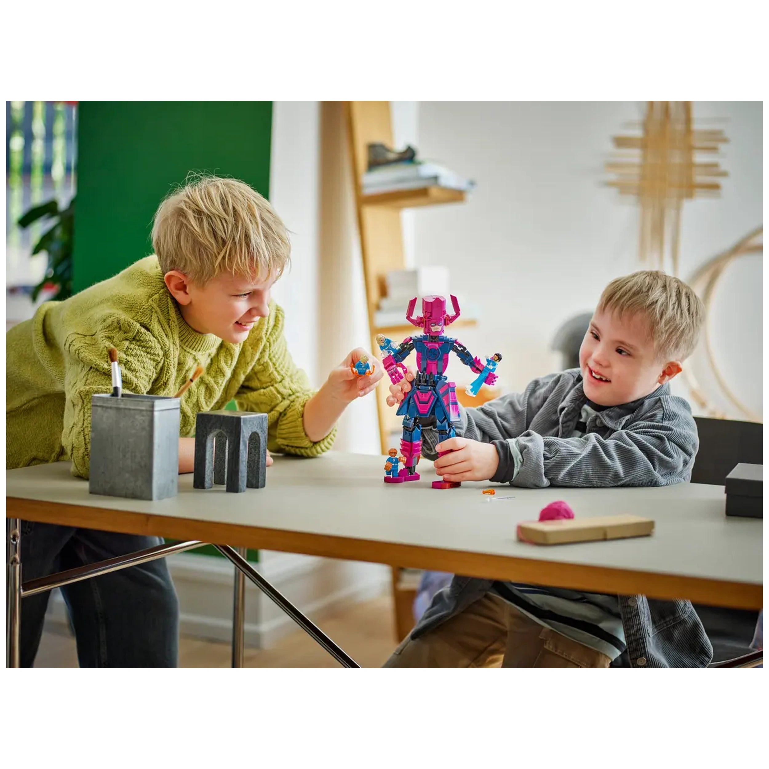 Two children play joyfully at a table, interacting with a colorful LEGO construction set featuring a large pink robot. One child, wearing a green sweater, holds a small piece while the other, in a gray jacket, smiles, showing excitement as they engage with the toy. The setting includes simple gray and wood decor, enhancing the playful atmosphere.