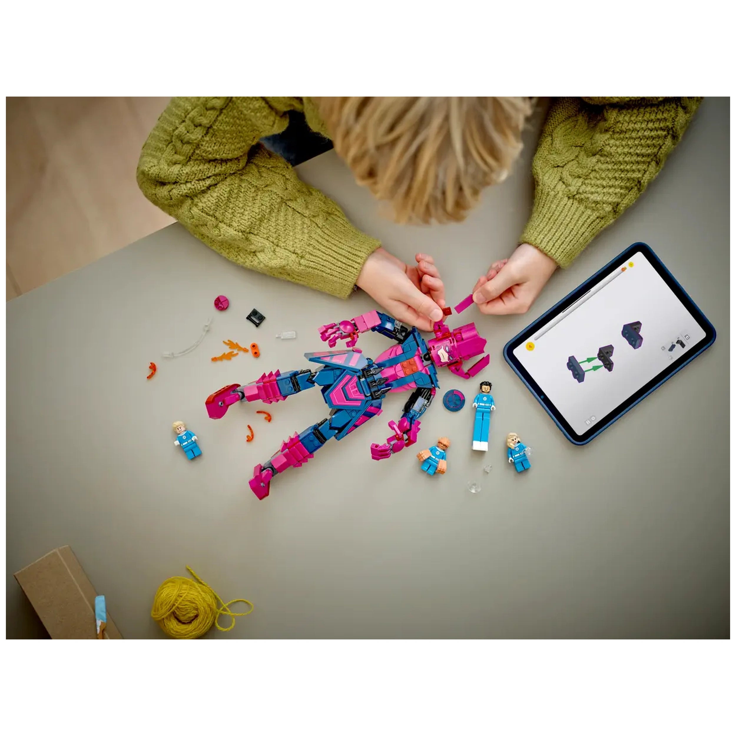 Child assembling a pink and blue LEGO figure with tiny parts scattered around, while following instructions on a tablet.