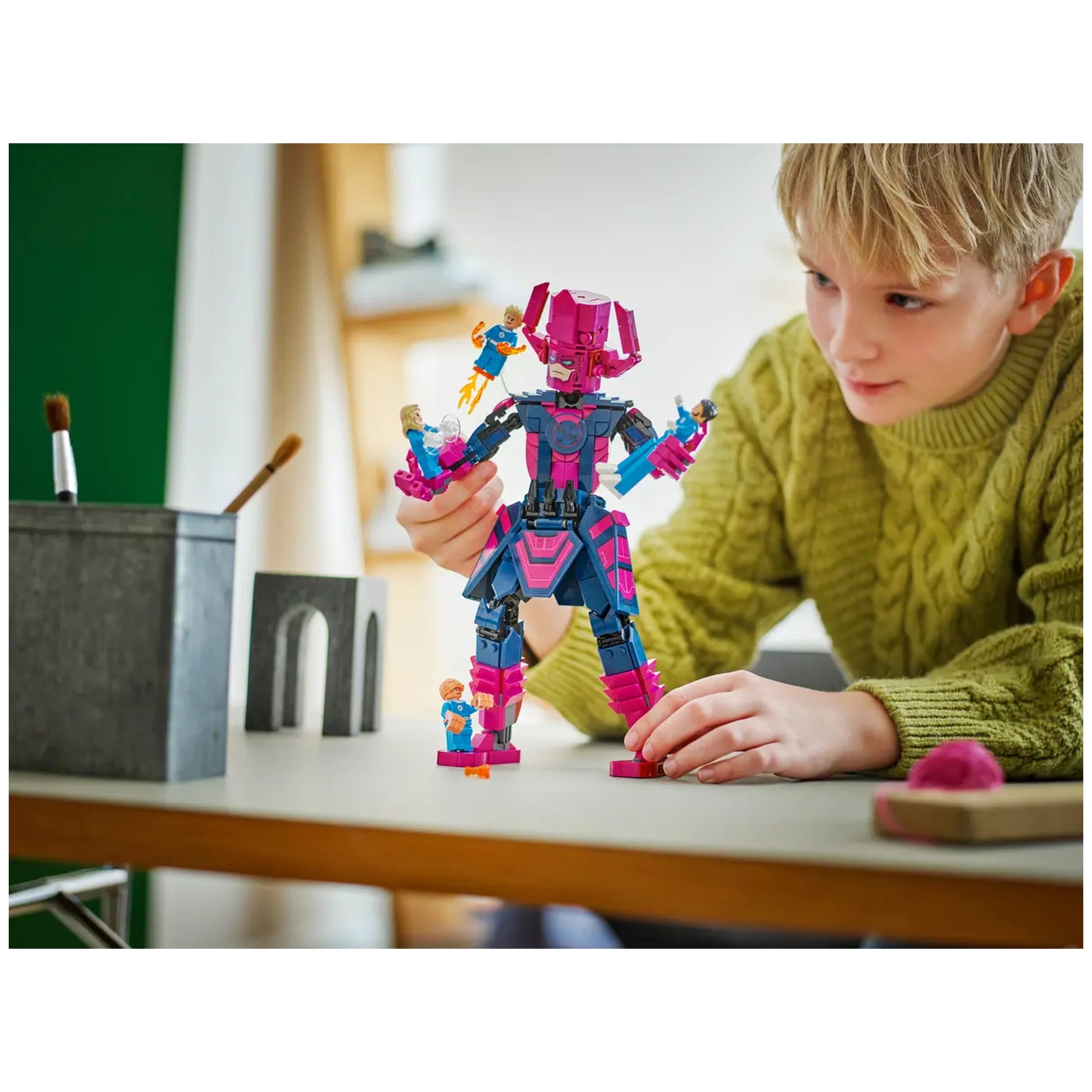 A child wearing a green sweater intently assembles a colorful LEGO figure resembling a superhero, surrounded by smaller figures and creative tools on a table.
