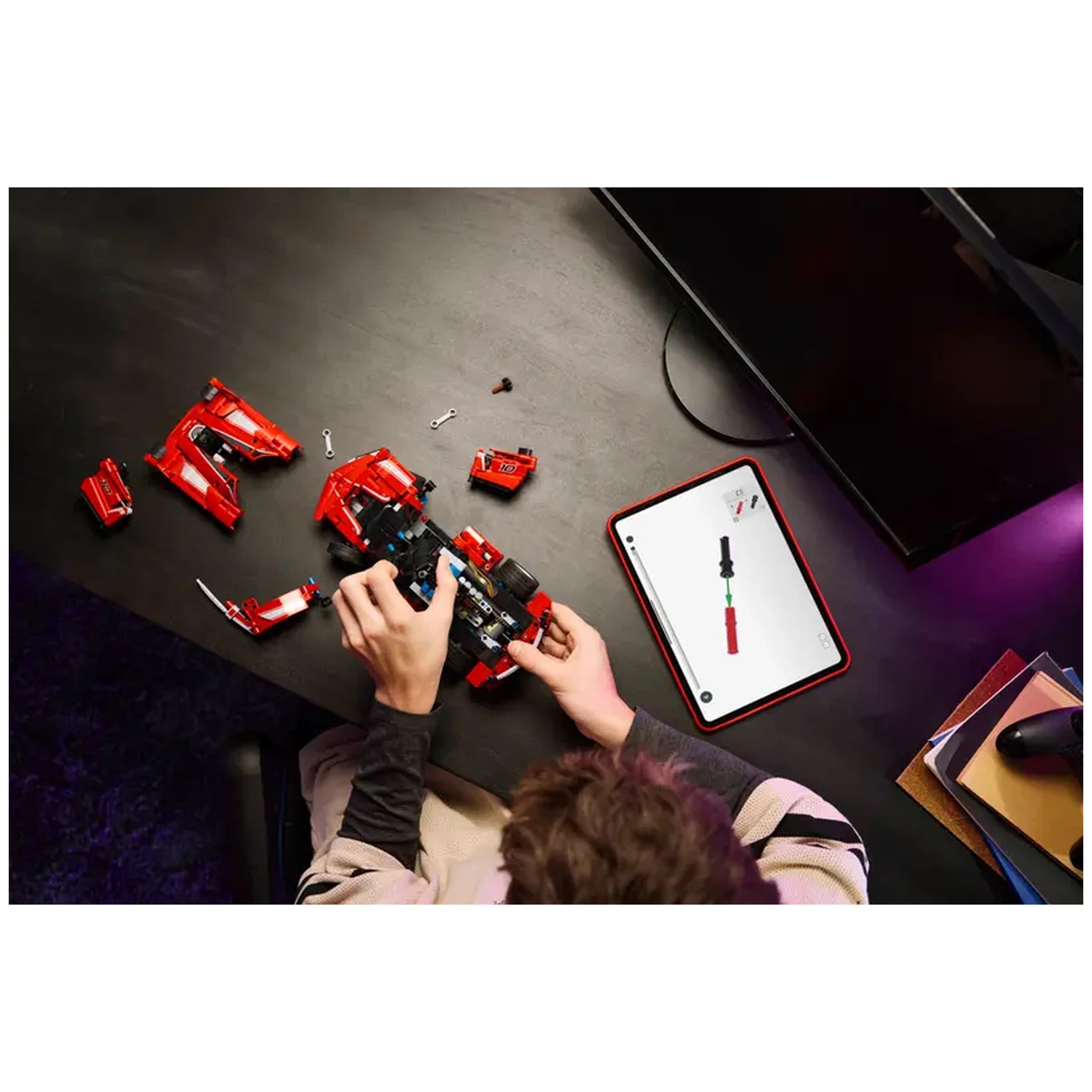 A person assembling a red LEGO Ferrari FXX K model kit on a dark table, surrounded by loose pieces and a tablet displaying assembly instructions.