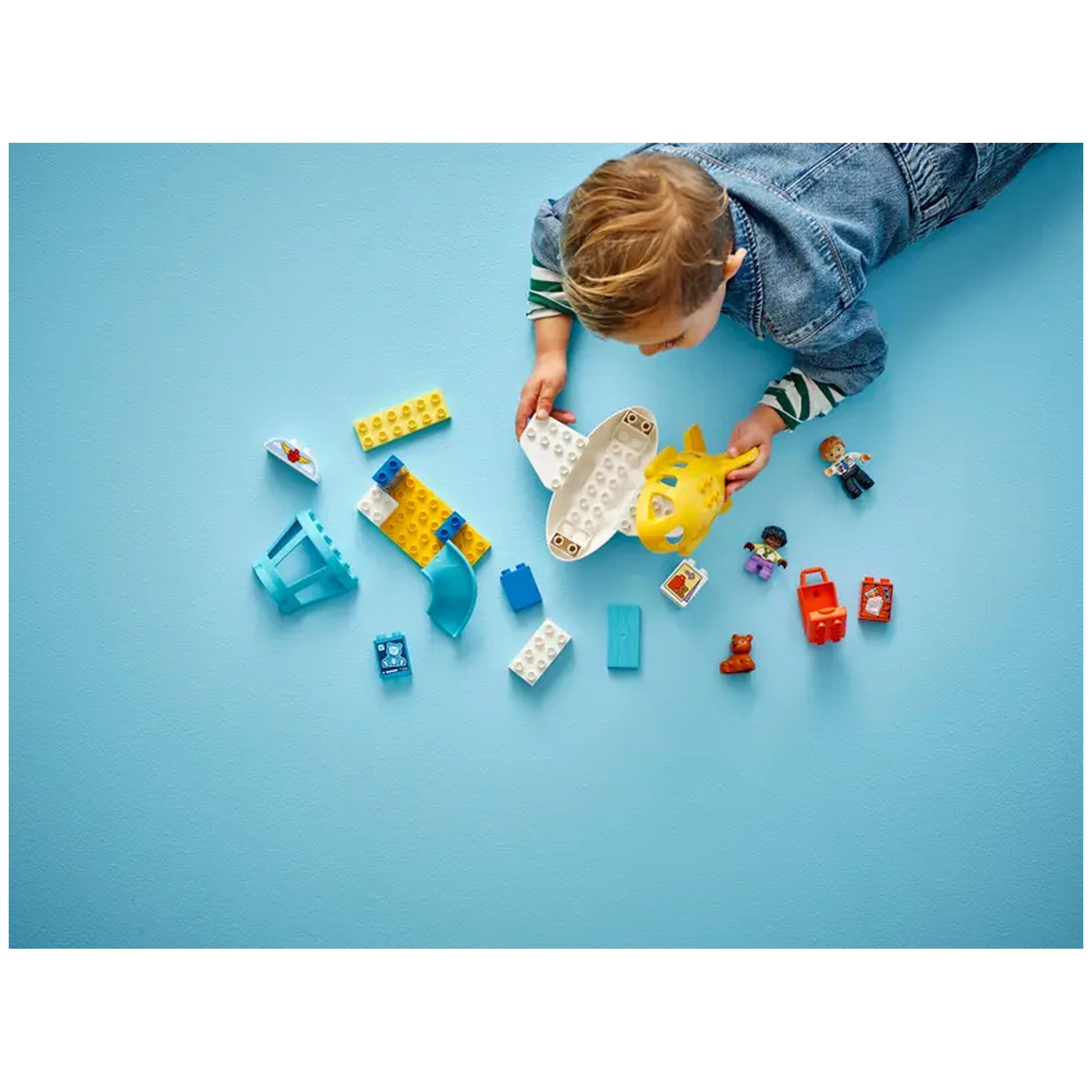A child lays on a light blue surface, joyfully assembling a LEGO airplane toy. Surrounding them are colorful building blocks, toy figures, and travel-themed accessories. The vibrant pieces include blue, yellow, and white elements, enhancing imaginative play.