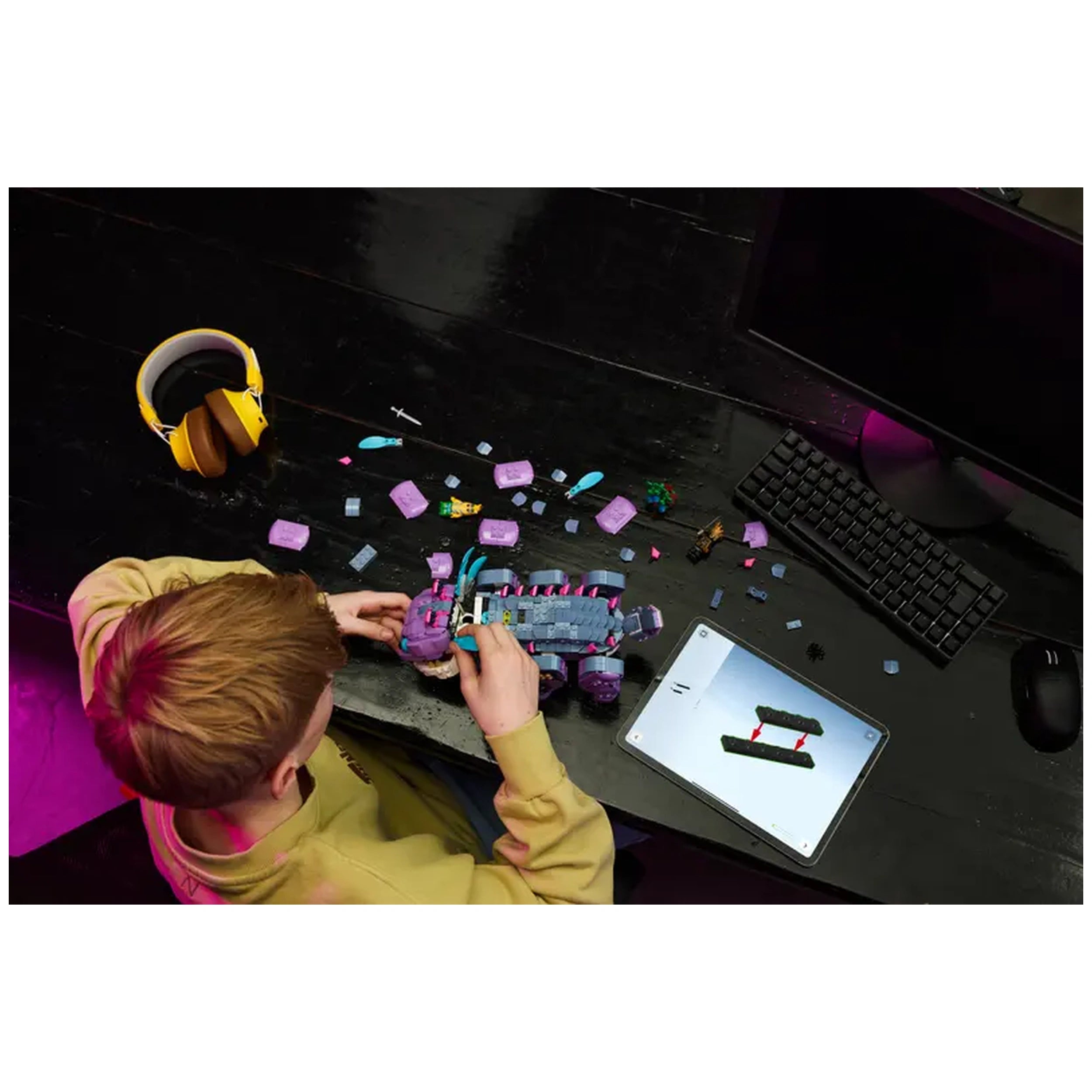 A child assembles the LEGO Fortnite Klombo set, surrounded by colorful pieces on a black table, with a tablet showing building instructions and headphones nearby.