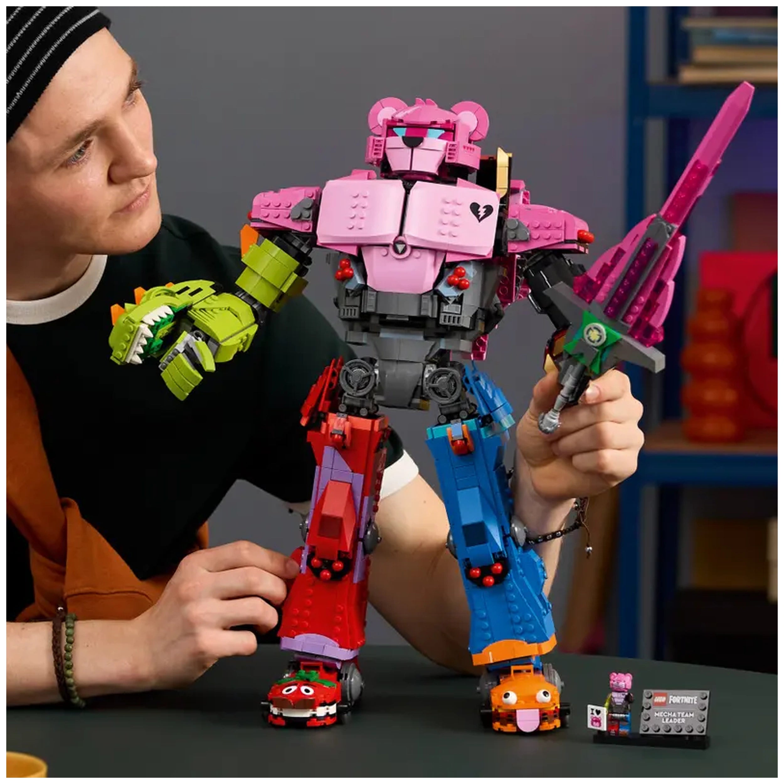 A young man holds the colorful LEGO Fortnite Mecha Team Leader figure, showcasing its dynamic pose and detailed features. The robot, featuring pink, red, blue, and green colors, has articulated limbs and a sword accessory. Near the figure are playful cartoonish elements resembling small vehicles. The backdrop includes a modern workspace with books and decorative items, creating a vibrant atmosphere.