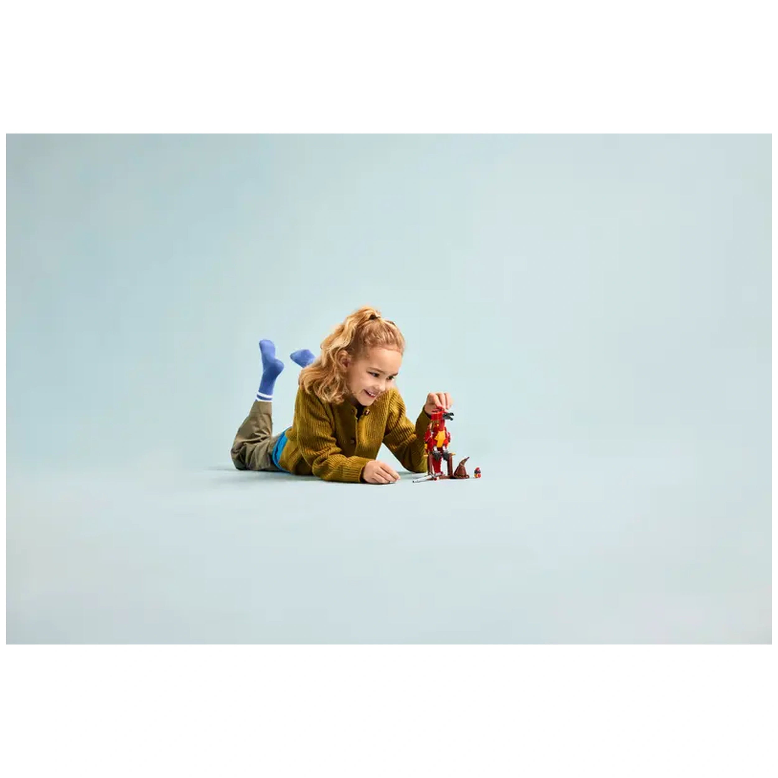 A joyful child with curly hair plays on the floor, examining a colorful LEGO® Fawkes figure alongside a buildable stand.