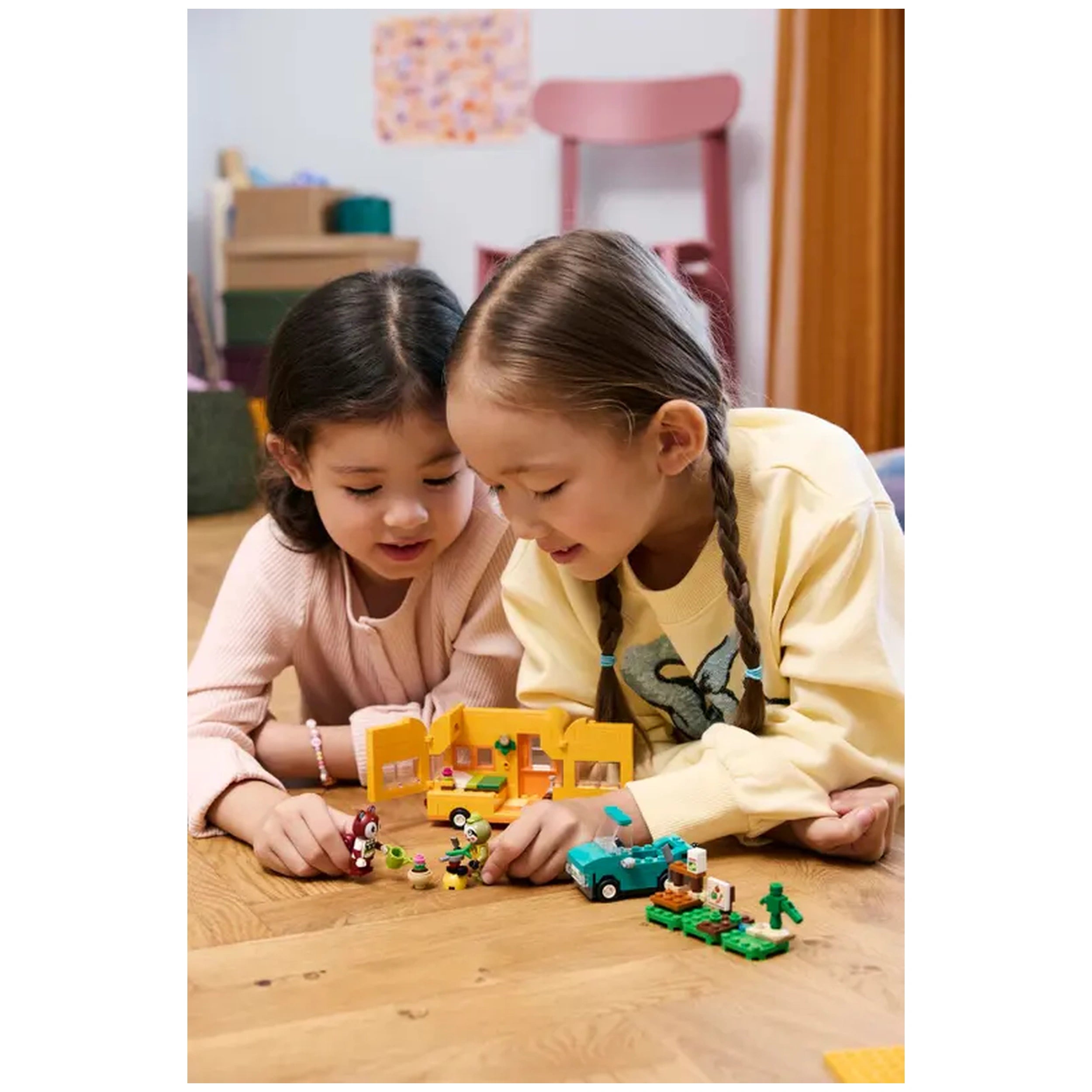 Two children play together on a wooden floor, engaging with the LEGO® Animal Crossing™ Leif's Caravan & Garden Shop set. Vibrant pieces, including a car and minifigures, are scattered around them. One child wears a light pink sweater and the other a yellow shirt with an animal design. They lean closely, showing excitement and creativity as they explore their imaginative play. In the background, soft colors and shapes of a cozy room are visible.