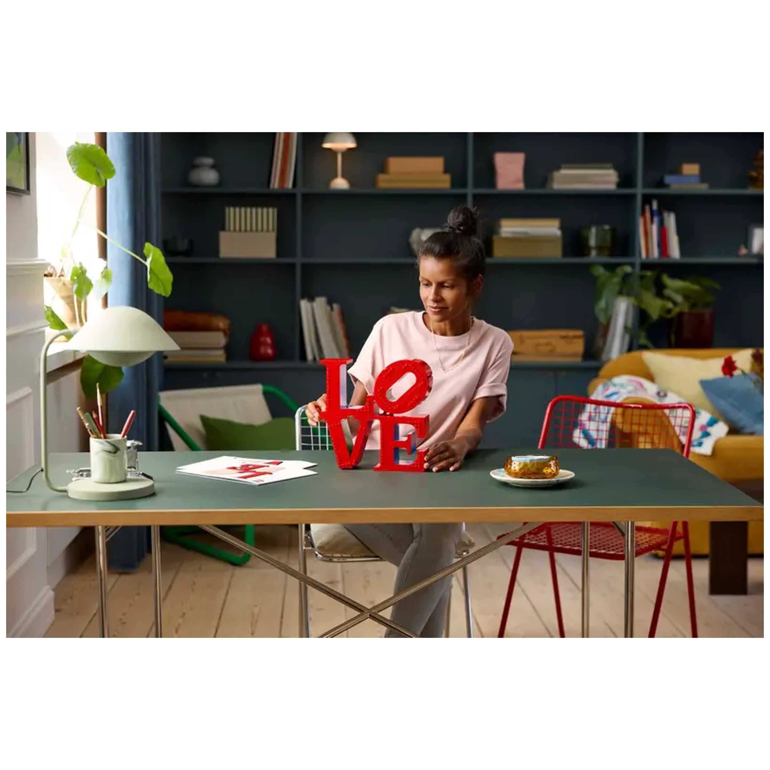 A woman placing a vibrant red LEGO® "LOVE" sculpture on a green table, surrounded by art prints, a dessert on a plate, and a stylish living room backdrop.