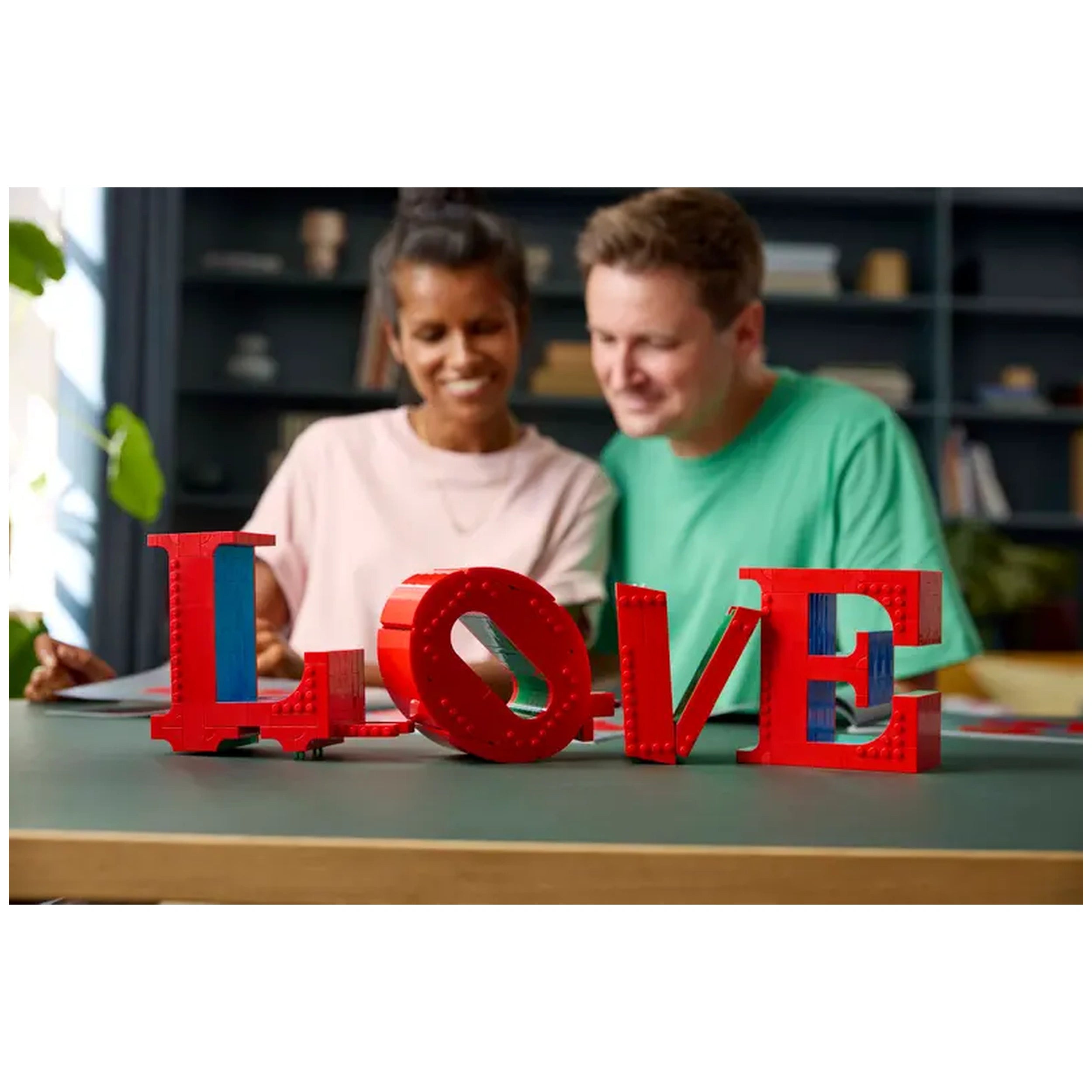 A red LEGO® construction set spelling "LOVE" sits on a table, while a smiling couple in casual attire engages in a creative project.
