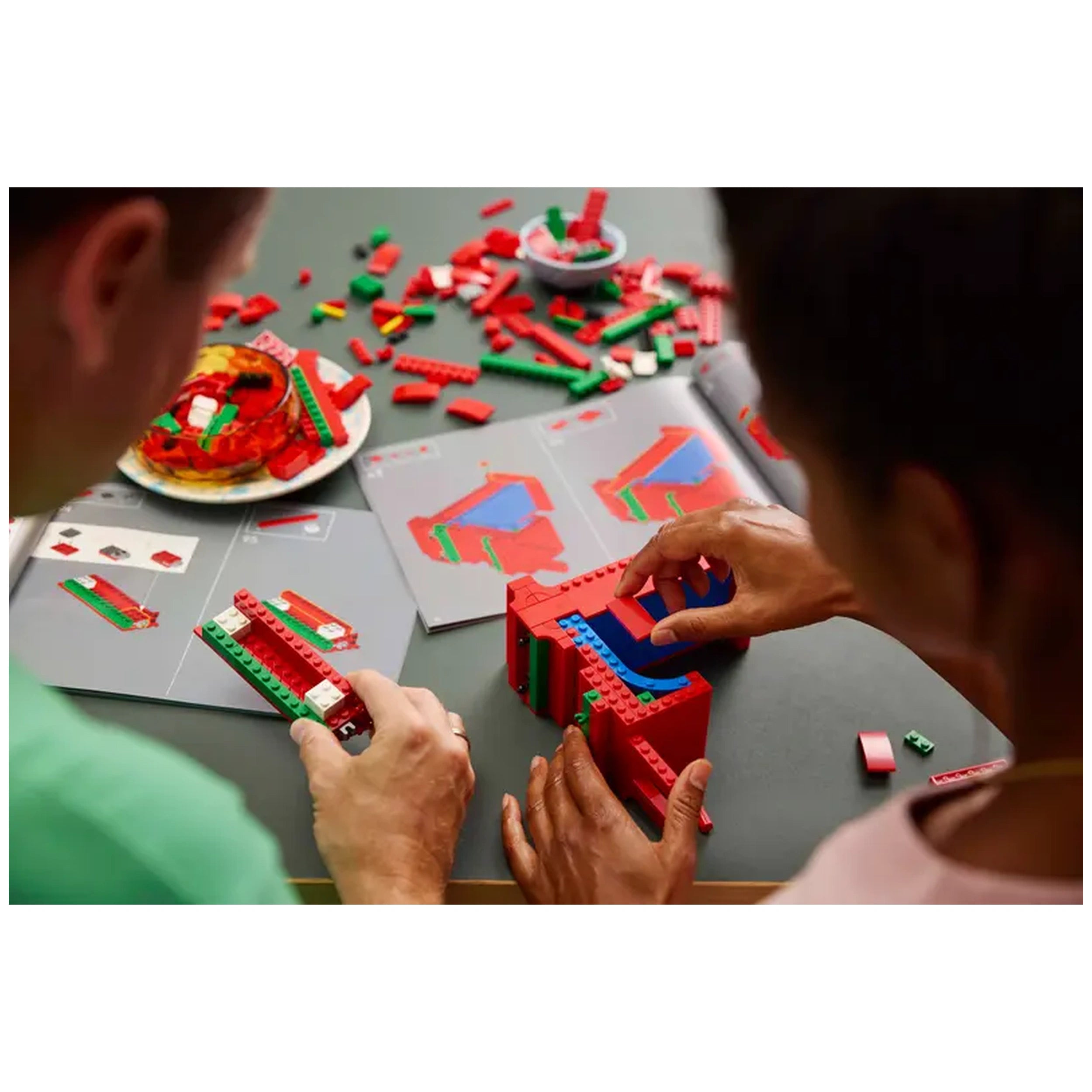 Two people collaborate on a LEGO® Art LOVE set, assembling vibrant red, blue, and green pieces. Instruction manuals and scattered bricks are visible on the table.