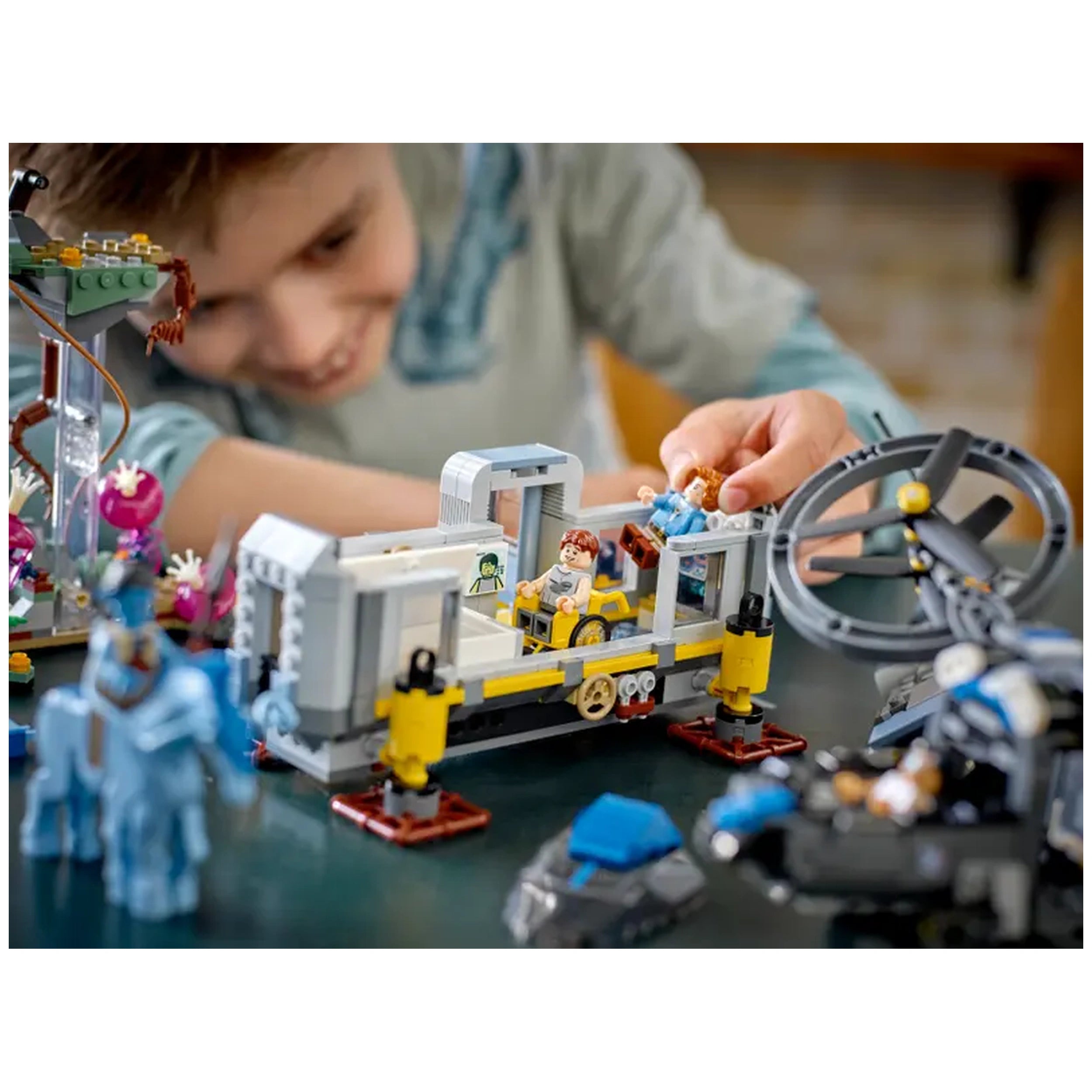 A young boy plays with the LEGO Avatar Floating Mountains set, featuring a gray container with two minifigures inside. He holds a Na'vi figure while focused on the colorful scene, which includes a blue Direhorse and a futuristic helicopter nearby.