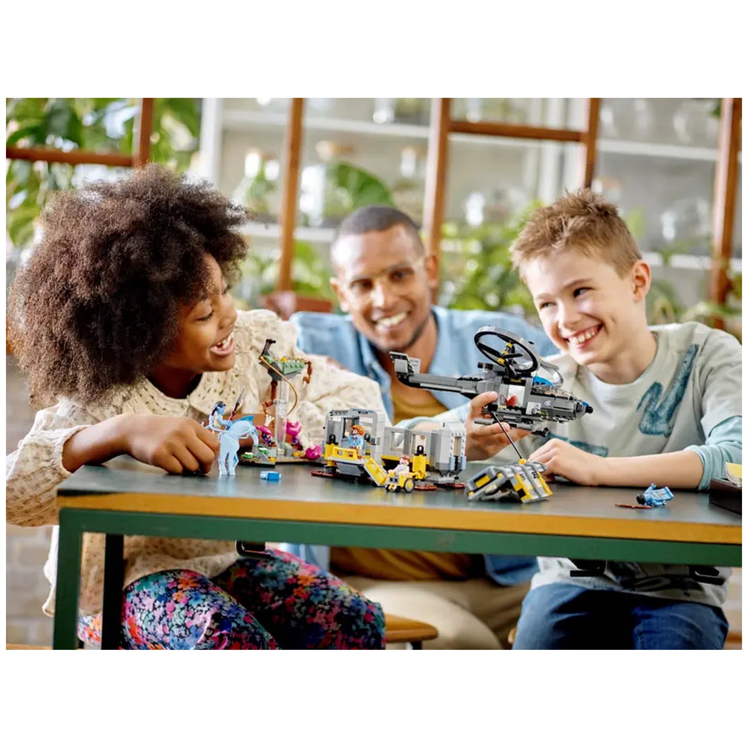 Two children joyfully play with the LEGO® Avatar Floating Mountains construction set on a green table, while a smiling adult watches them in a bright, plant-filled room. The set includes a futuristic helicopter, colorful minifigures, and various elements from the Avatar universe.