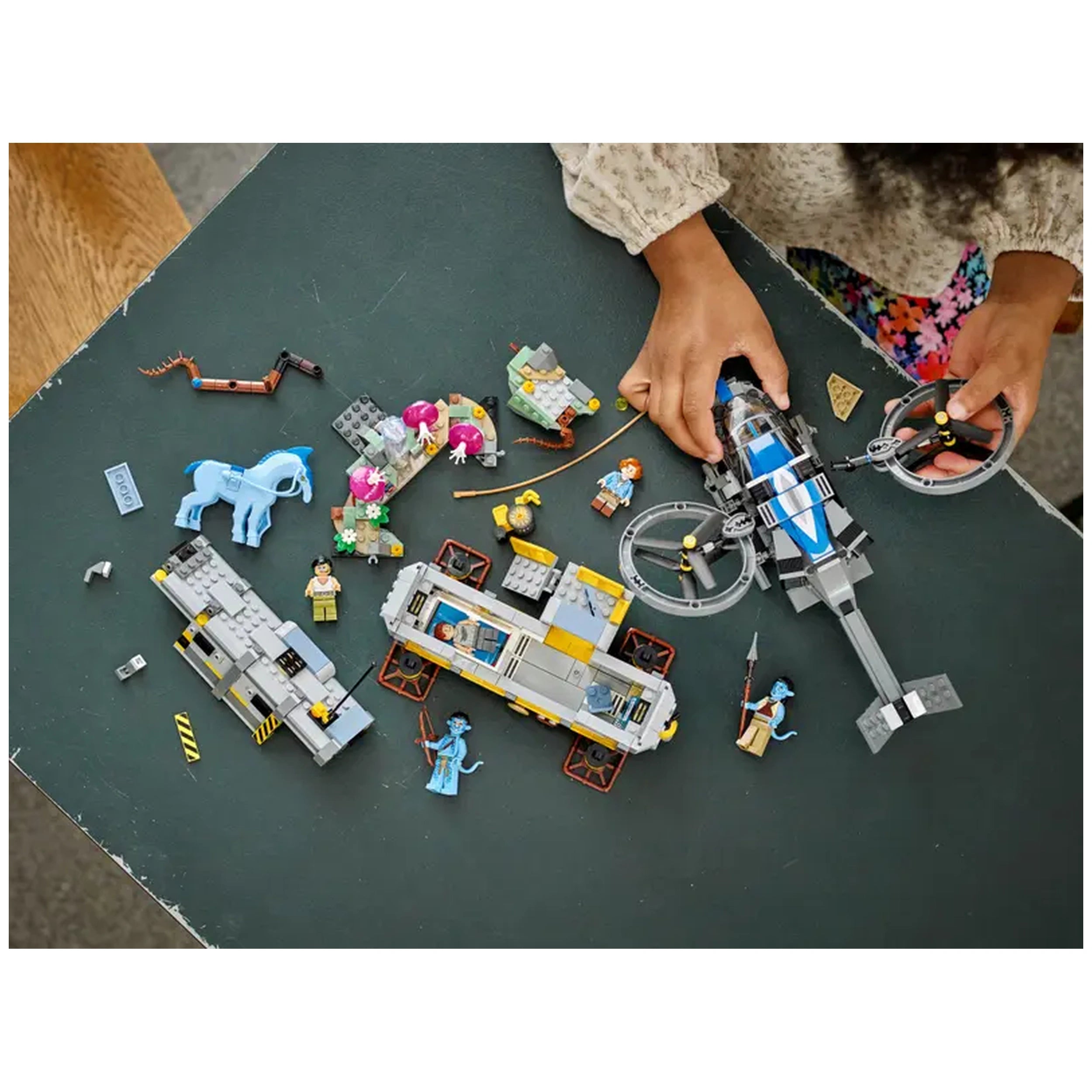 Child assembling the LEGO Avatar Floating Mountains set on a dark table, featuring colorful minifigures, a Direhorse, and a helicopter.