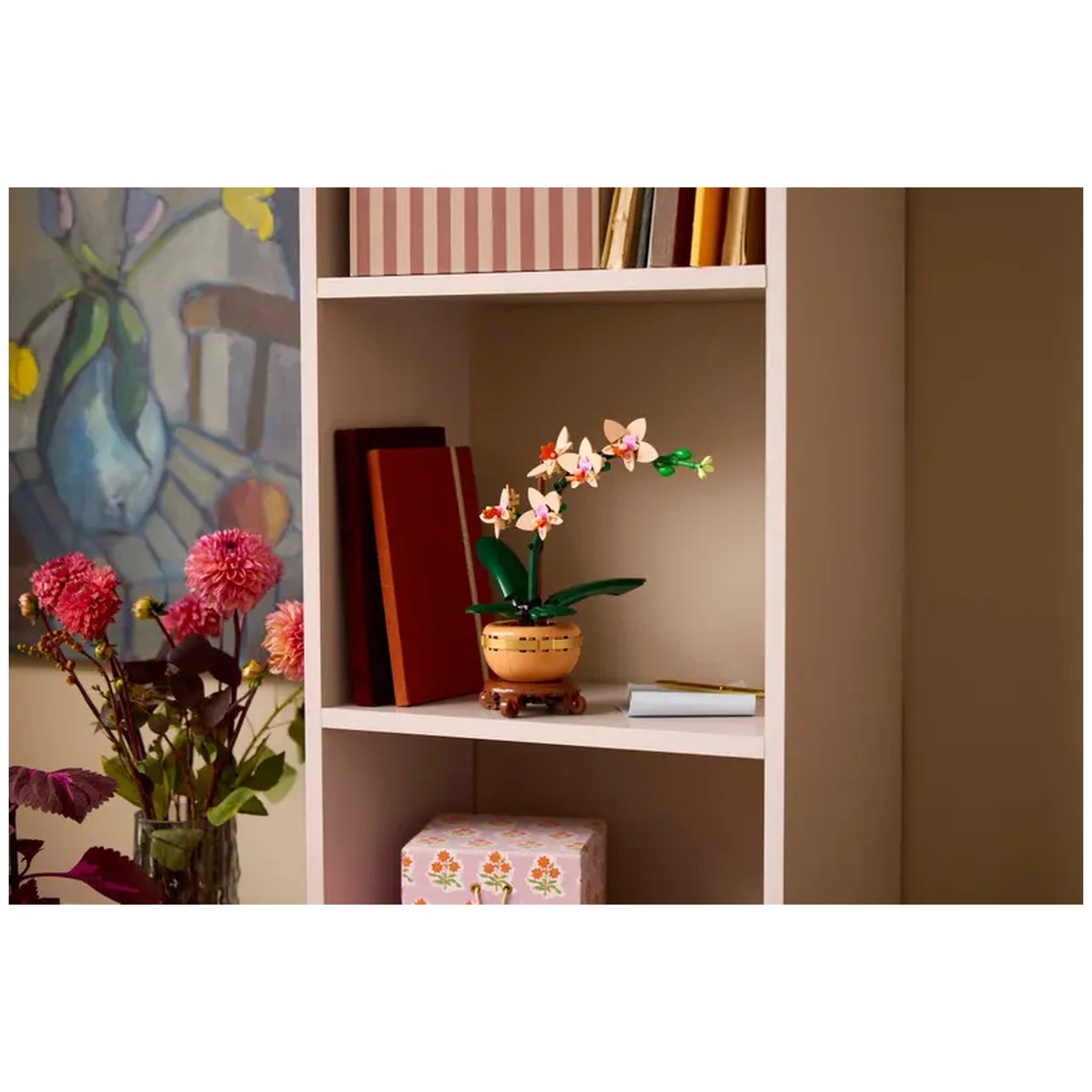 LEGO® Mini Orchid construction set displayed on a shelf, featuring peach blooms, green leaves, and a light terracotta pot, with colorful books in the background.