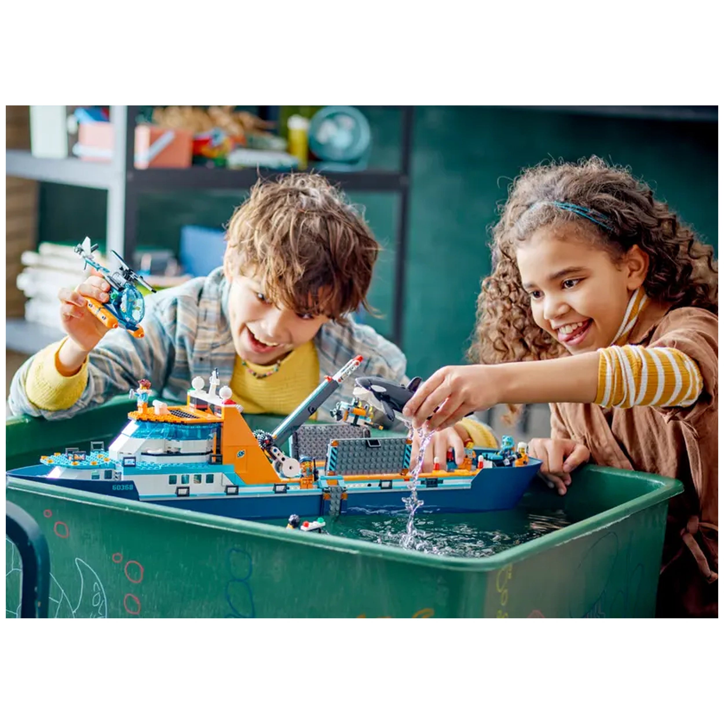 Two children joyfully playing with the LEGO® City Arctic Explorer Ship in a green tub filled with water, featuring a helicopter and colorful details.