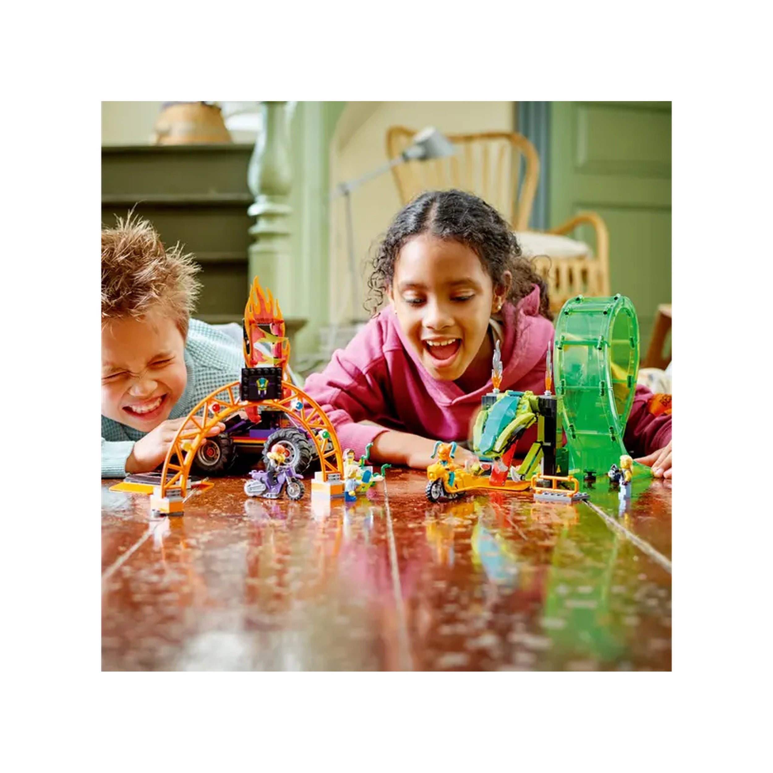 Two children play excitedly on a wooden floor with the LEGO City Double Loop Stunt Arena set, featuring colorful stunt bikes and props.