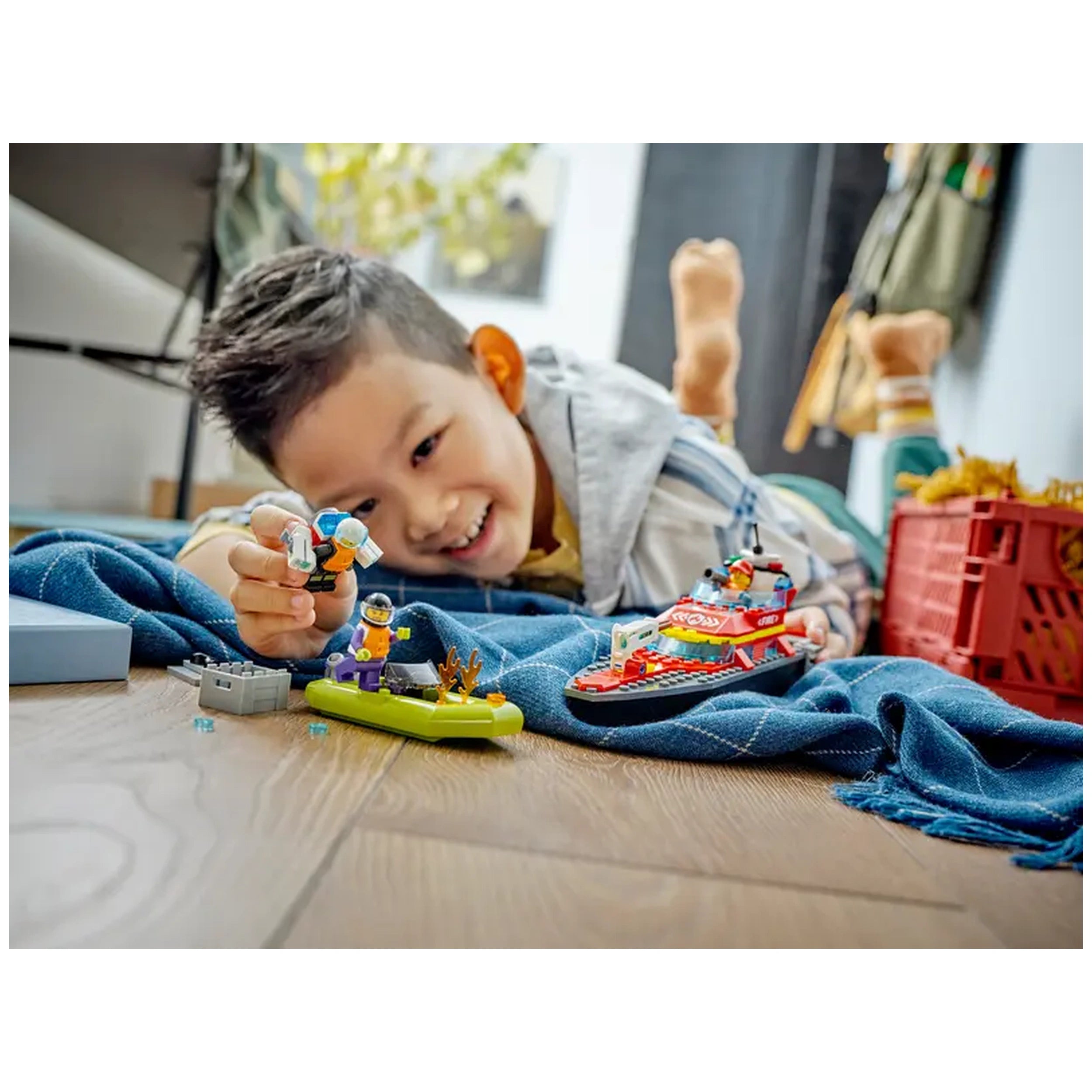 A young boy smiles while playing on a blue blanket, holding a LEGO firefighter minifigure. Nearby, a red toy fireboat and a green dinghy are positioned on the wooden floor, surrounded by small LEGO pieces.