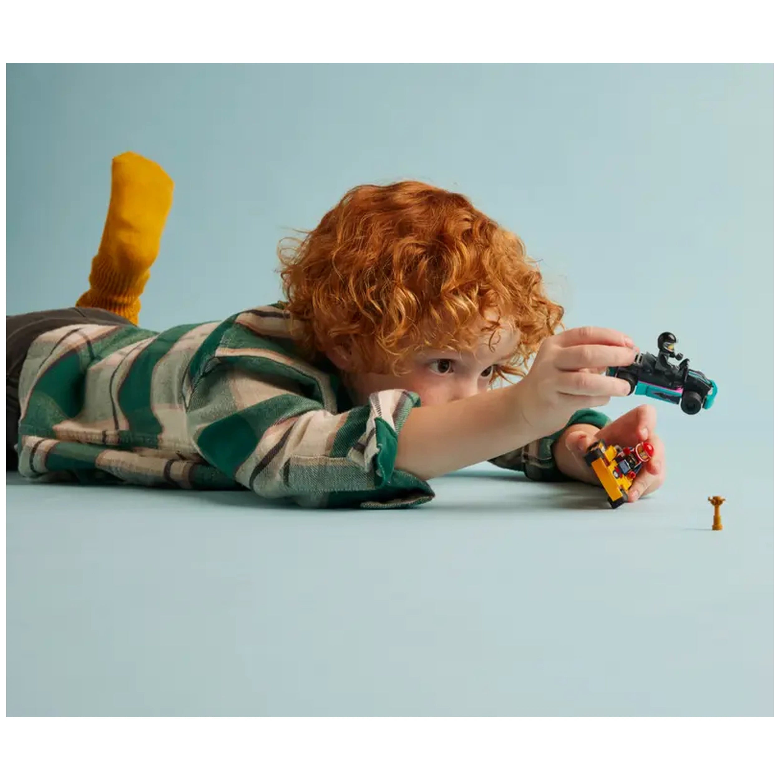 A young child with curly red hair lies on a light blue surface, focused on a LEGO construction set. They hold a mini go-kart in one hand and a driver minifigure in the other, with a trophy element nearby. The child wears a green and white checked shirt and bright yellow socks.