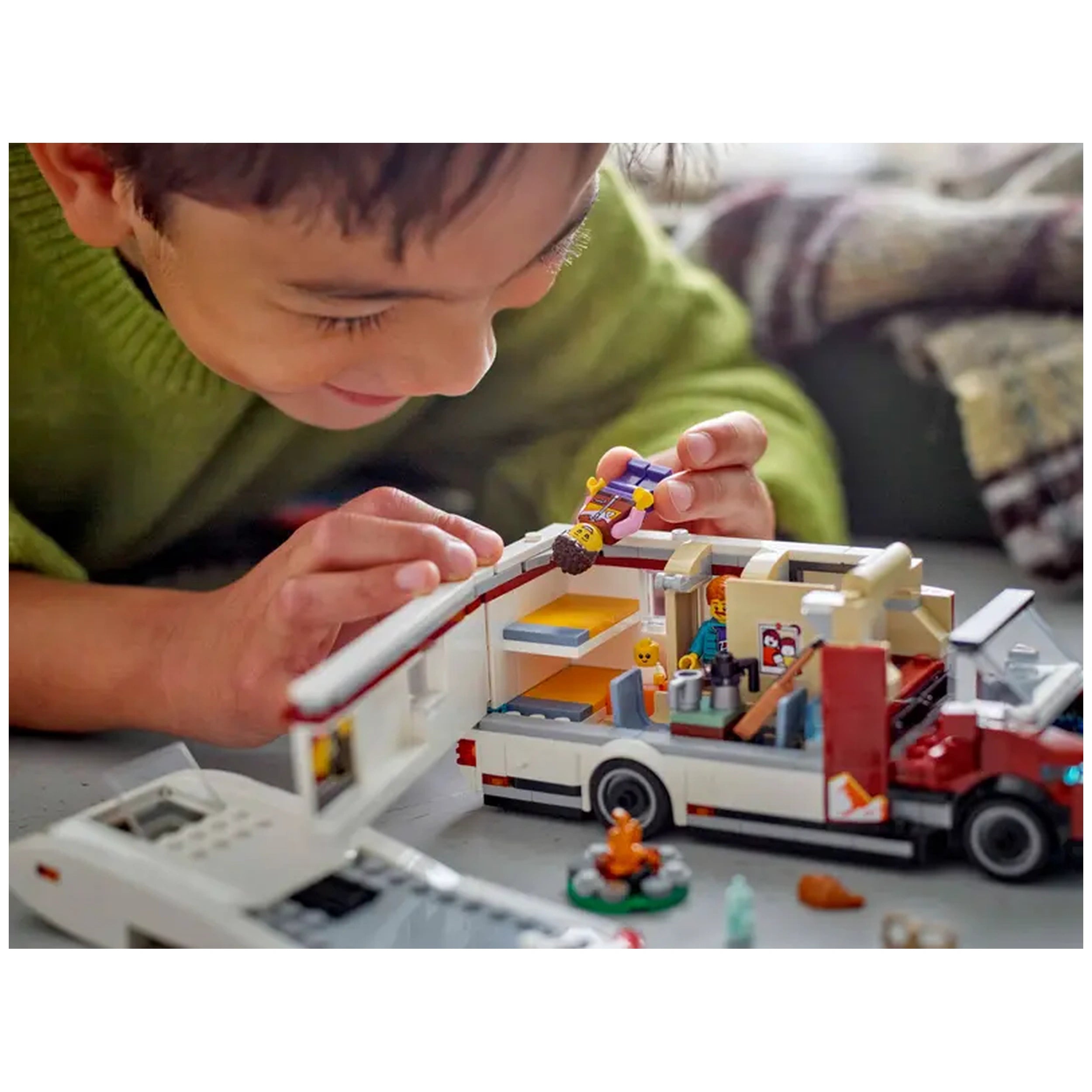A young child in a green sweater engages with a LEGO City Holiday Camper Van set, examining a minifigure while surrounded by vibrant interior details and fun accessories like a toy campfire.