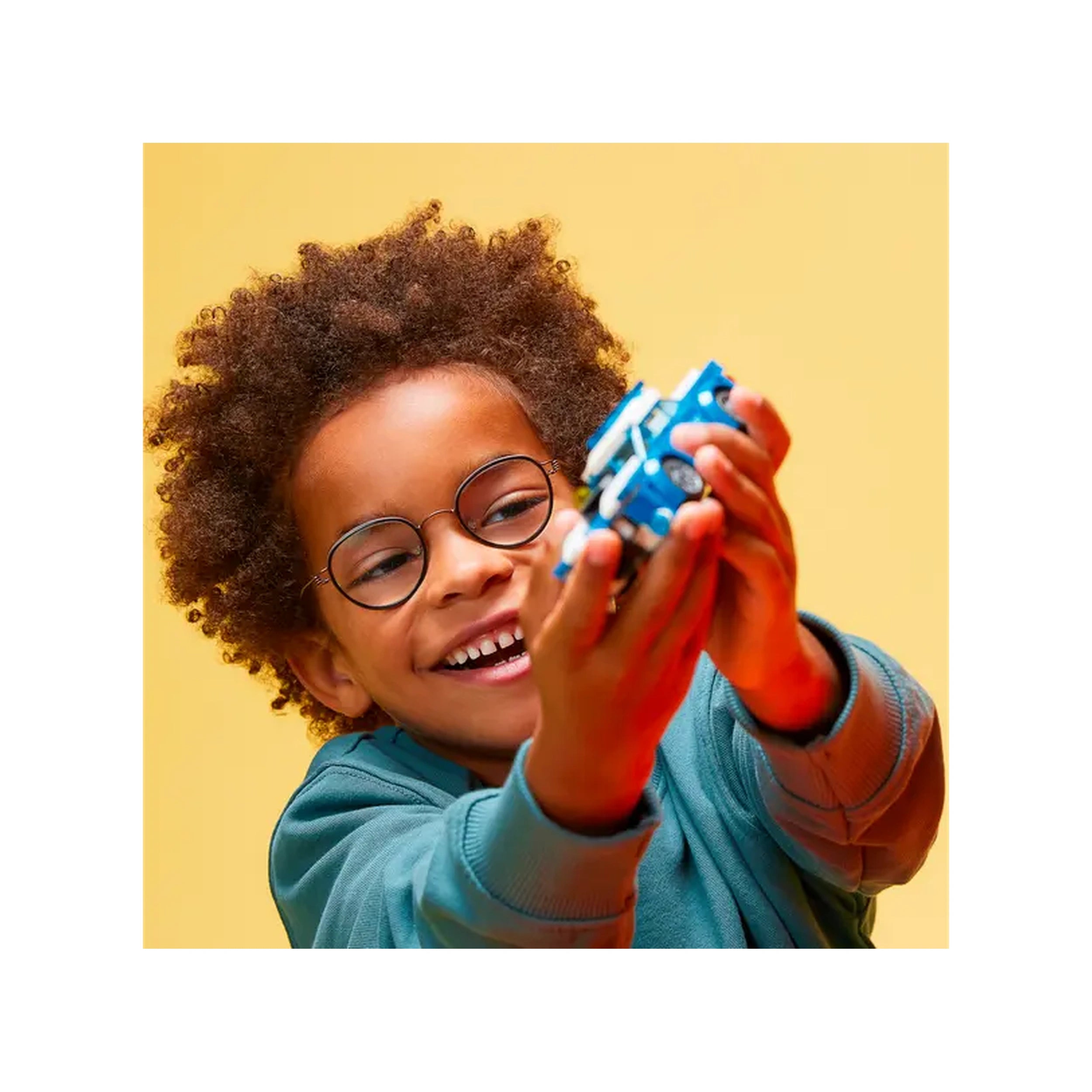 A smiling child with curly hair and glasses joyfully holds a blue LEGO police car, set against a bright yellow background.
