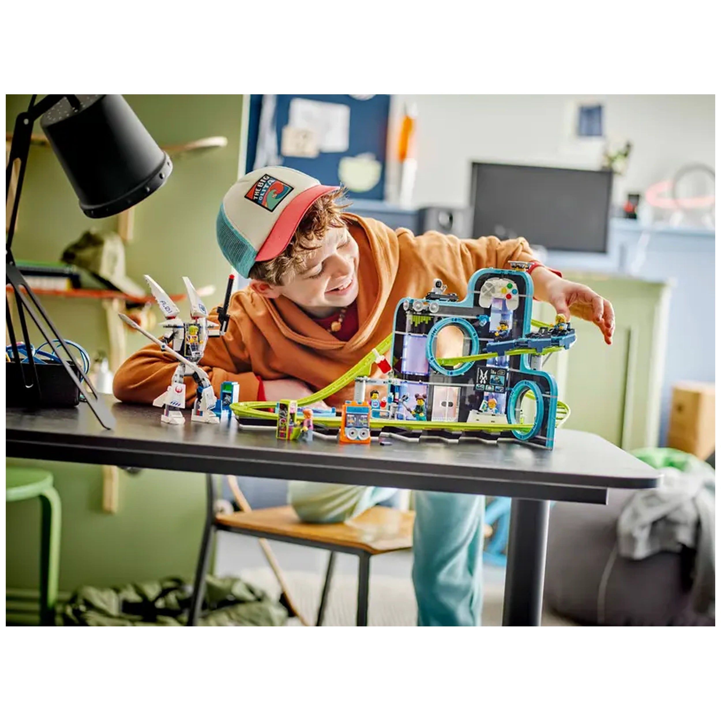 A child with curly hair and a cap smiles while playing with a colorful LEGO® City Robot World Roller-Coaster Park set on a table. The set features a vibrant roller coaster, arcade elements, and an articulated mech suit. A lamp and wires are visible in the background, adding to the playful atmosphere of the room.