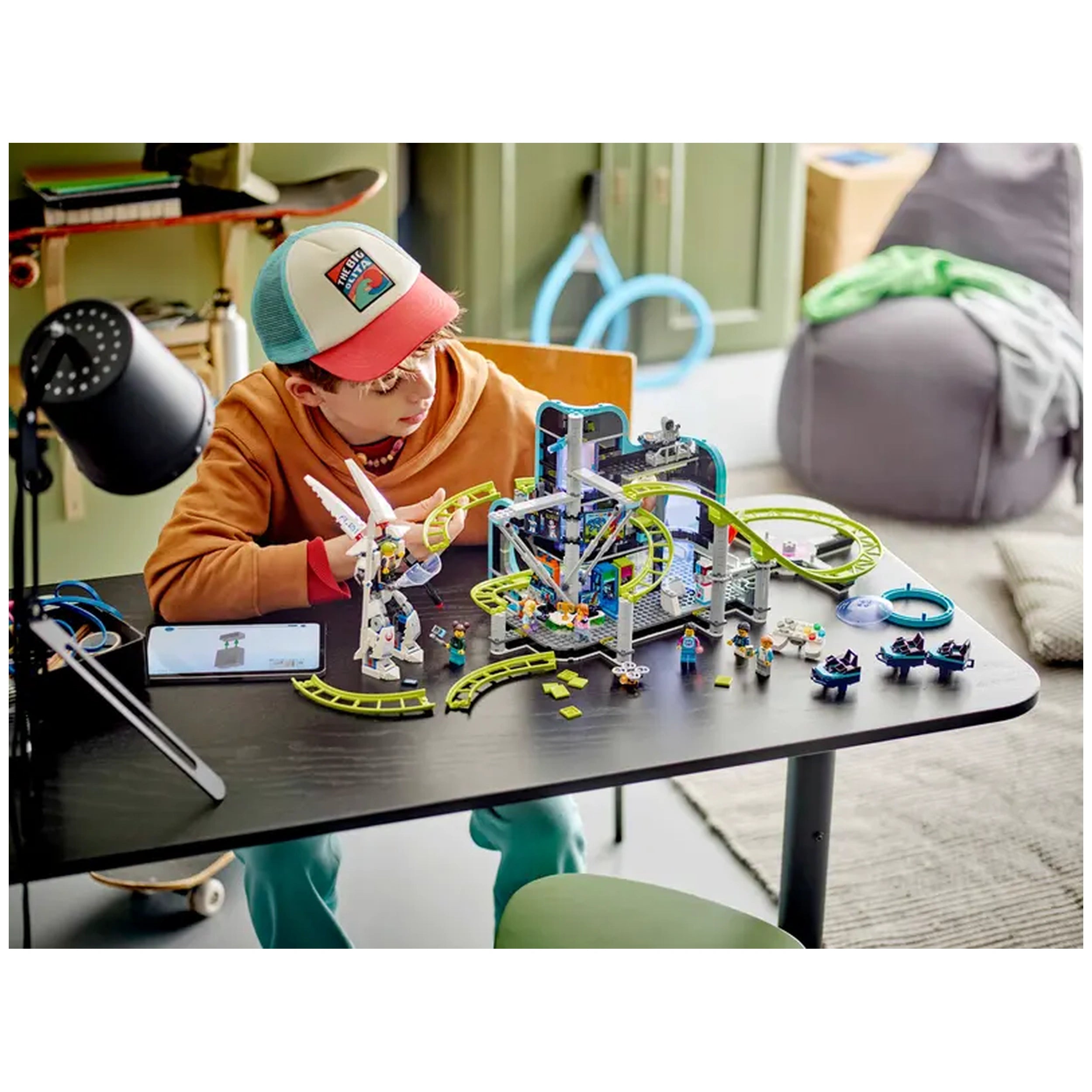 A child with a red and blue cap sits at a black table, focused on a colorful LEGO® City Robot World Roller-Coaster Park set, featuring tracks, a futuristic arcade, and playful characters. A smartphone lies beside them, and the background includes soft furnishings.