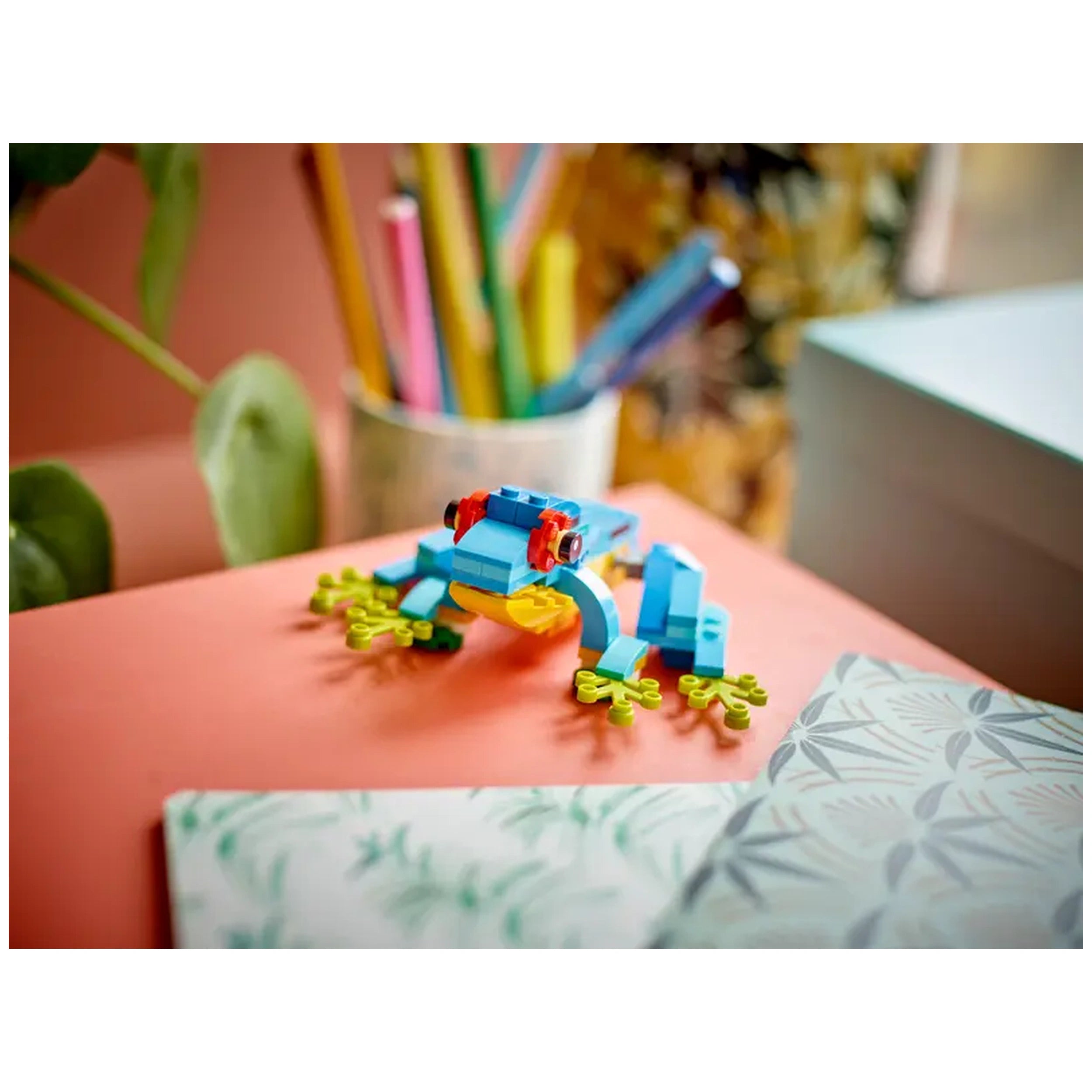 Colorful LEGO® Creator frog in blue and green, with a yellow belly, sitting on a vibrant coral surface, surrounded by plants and colored pencils.