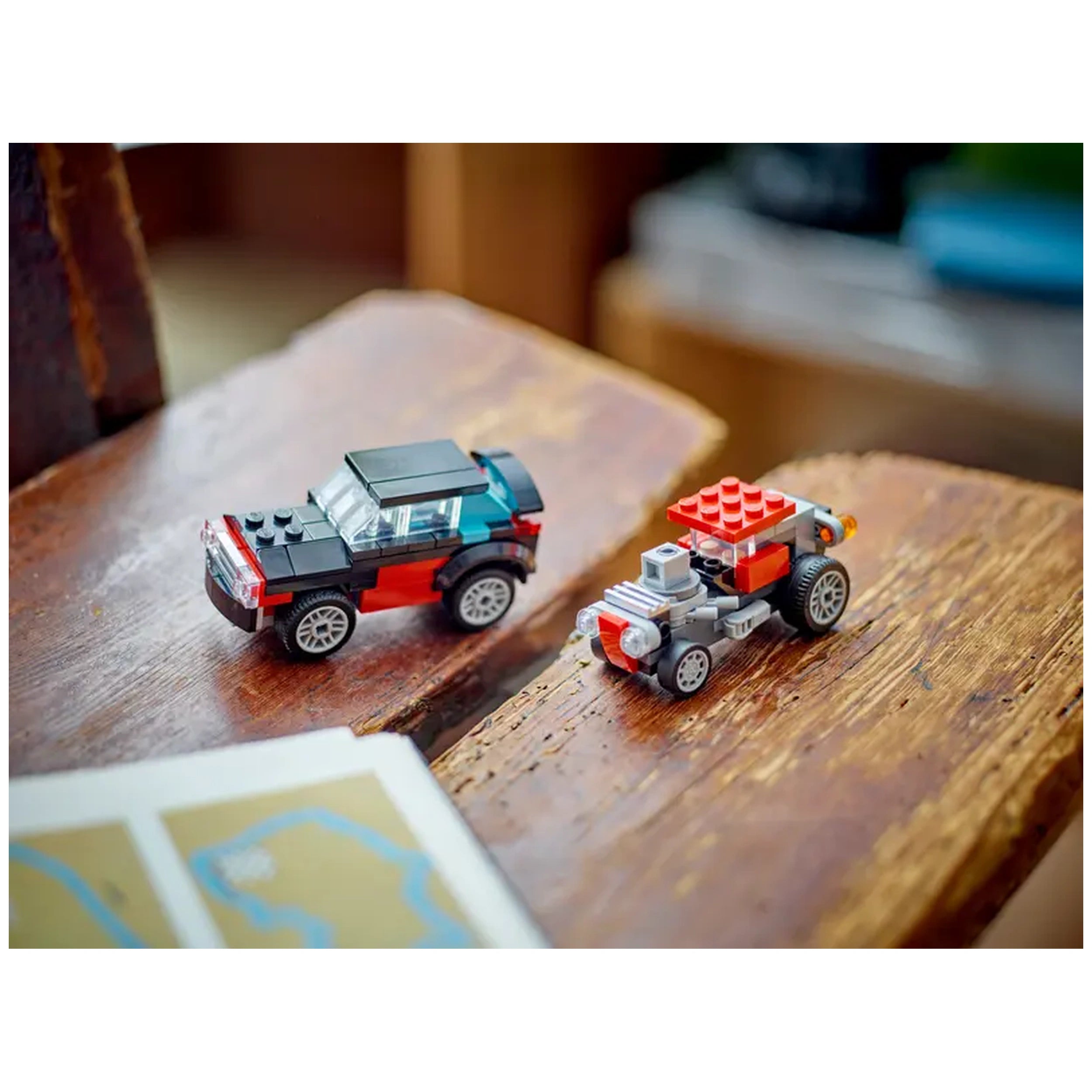 Two LEGO cars on a wooden surface: one sleek black and red vehicle with a clear windshield, and a smaller red and gray car with a prominent engine detailing. A map is partially visible in the foreground.