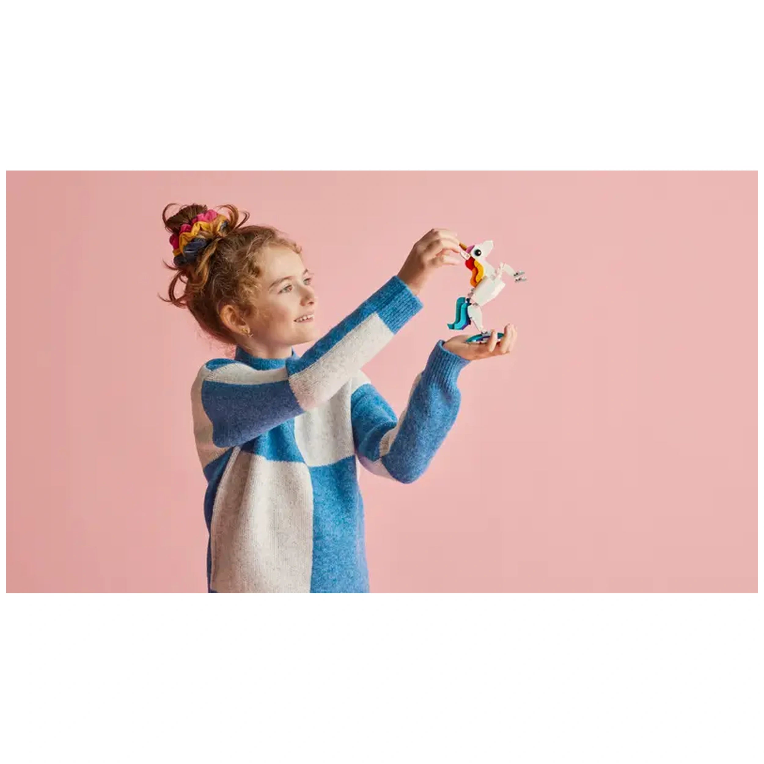 A smiling child with curly hair holds a colorful LEGO Creator Magical Unicorn, featuring a golden horn and vibrant tail, against a pink background.