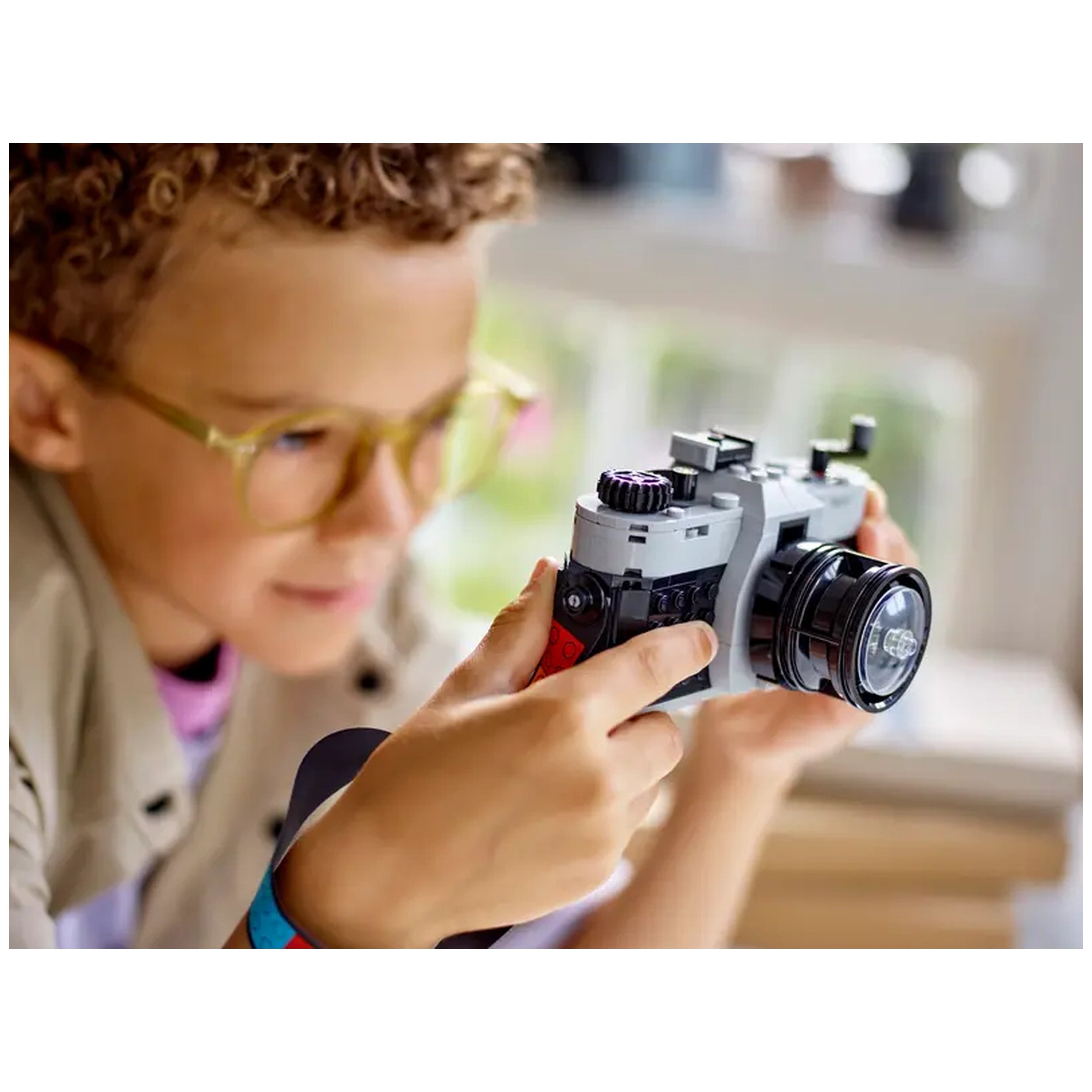 A young child with curly hair and yellow glasses holds a LEGO® Creator Retro Camera, focused on play. The camera features a sleek design with a black lens and moving parts, set against a bright background.