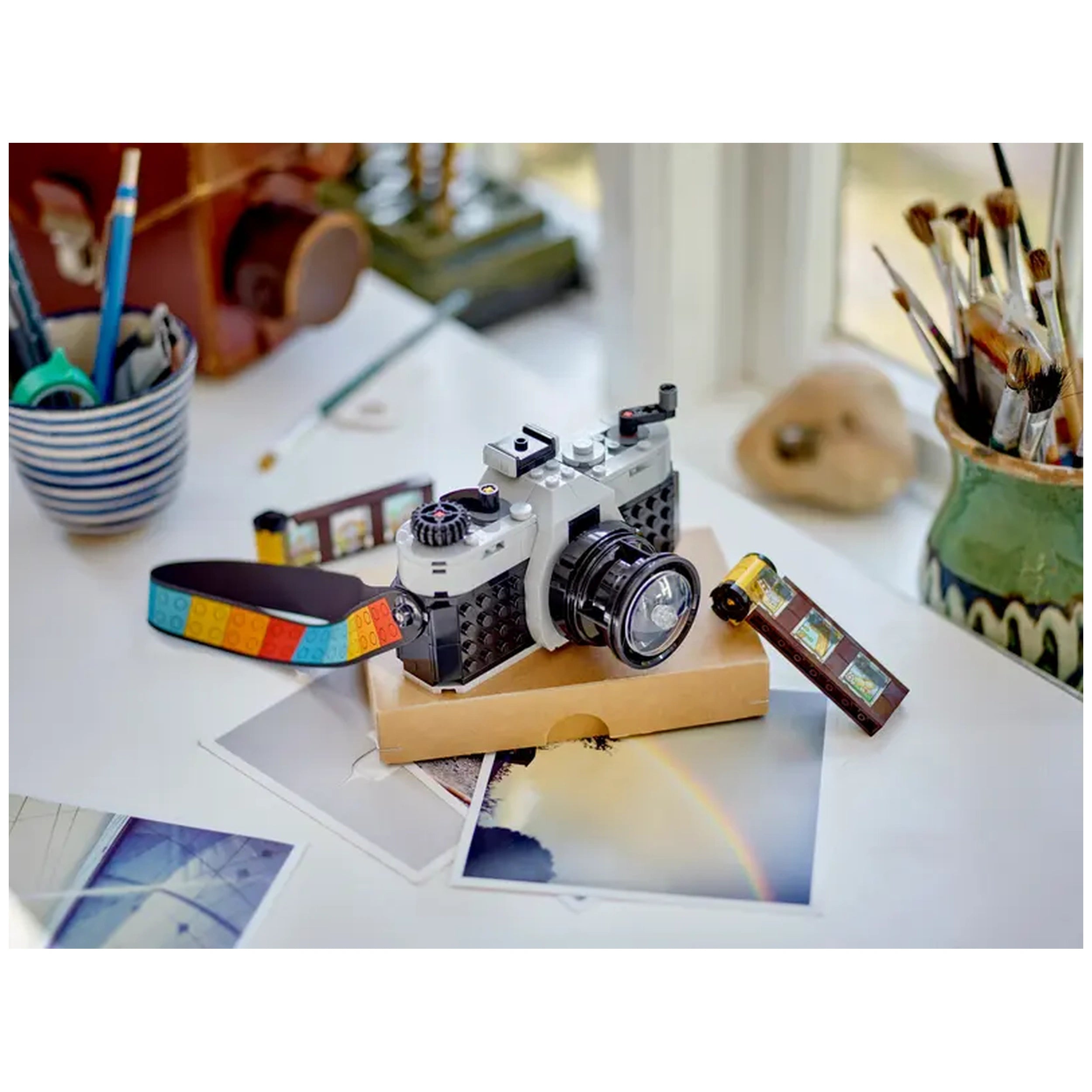 LEGO® Creator Retro Camera set displayed on a table, surrounded by printed photos. The camera features a retro design with a colorful strap and a brick-built film piece.
