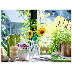 Two vibrant LEGO sunflower blooms in a clear bottle, surrounded by a decorative jug, green watering can, and potted plants, on a sunlit table.