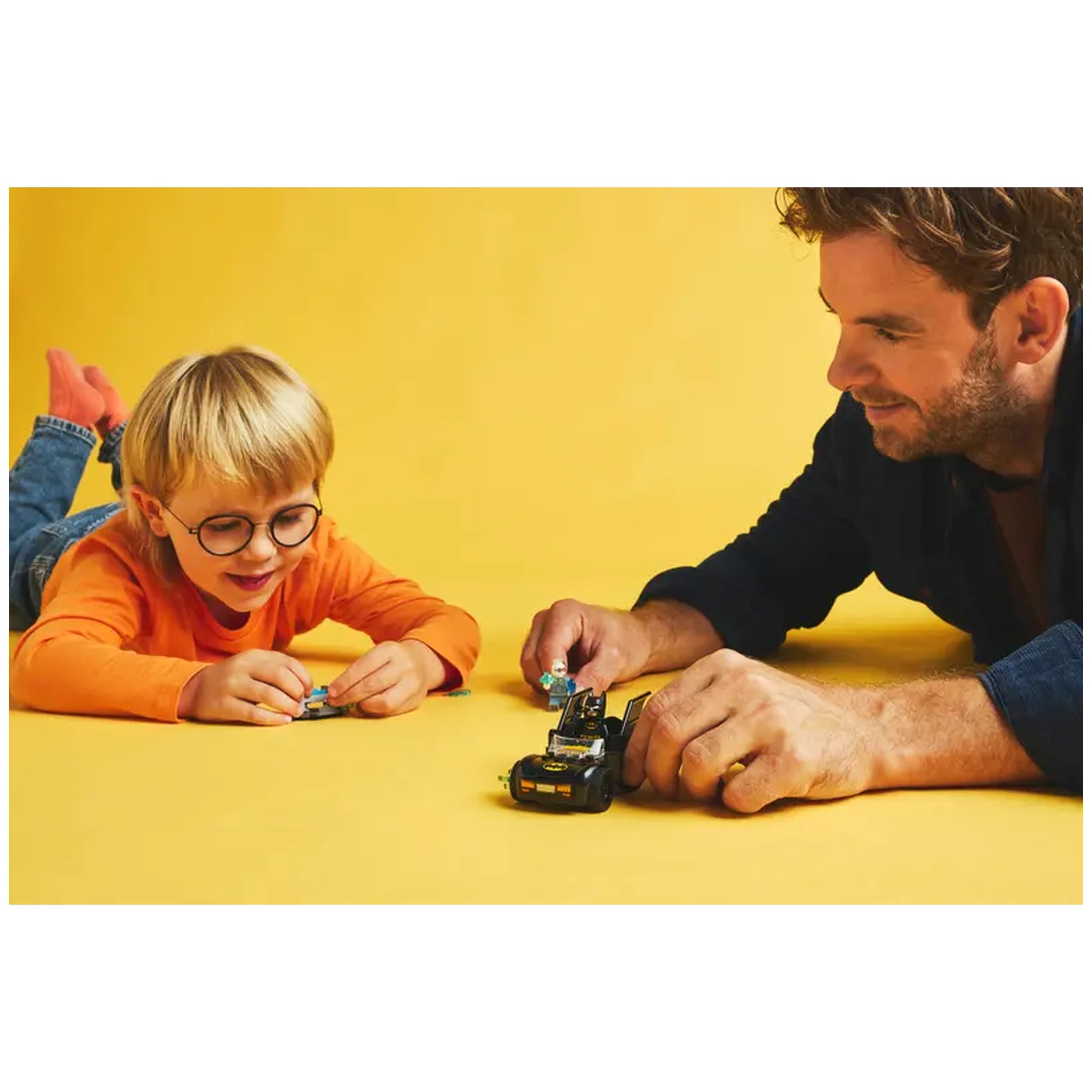 A young boy in an orange shirt and glasses smiles while playing with LEGO pieces alongside an adult man, both focused on a black Batmobile set against a bright yellow background.