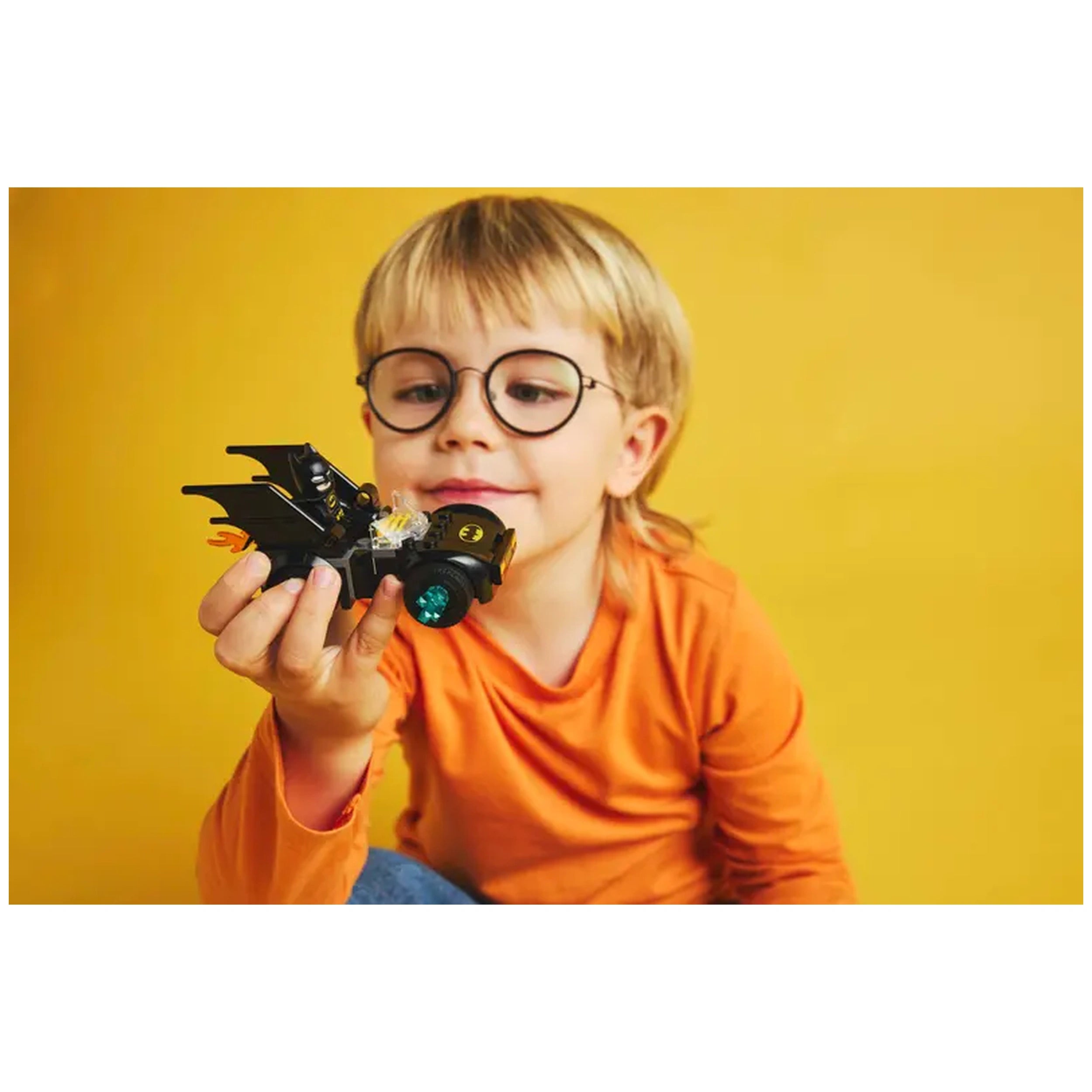 A young child with glasses holds a black Batmobile toy against a bright yellow background, smiling with excitement.