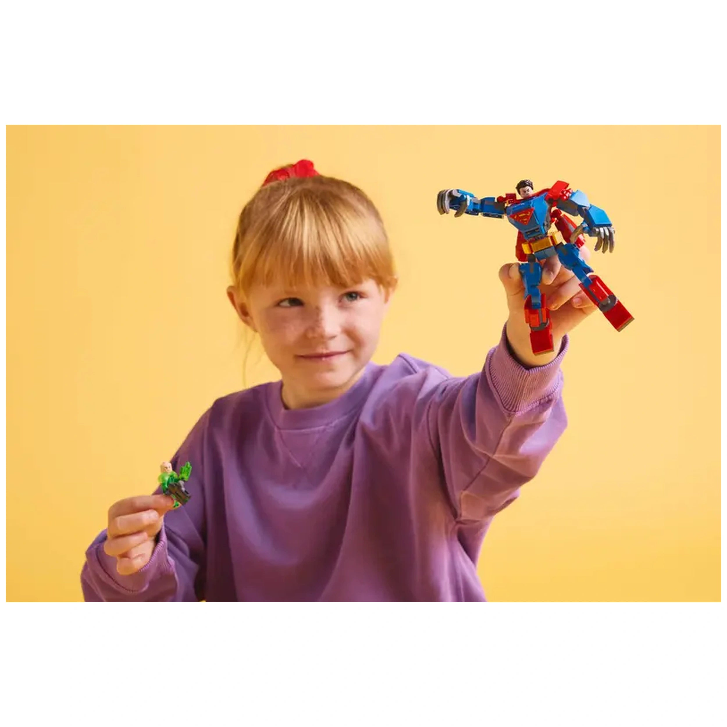 A young girl in a purple sweatshirt joyfully holds a bright red and blue Superman Mech toy in one hand and a green mini-figure in the other, set against a vibrant yellow background.