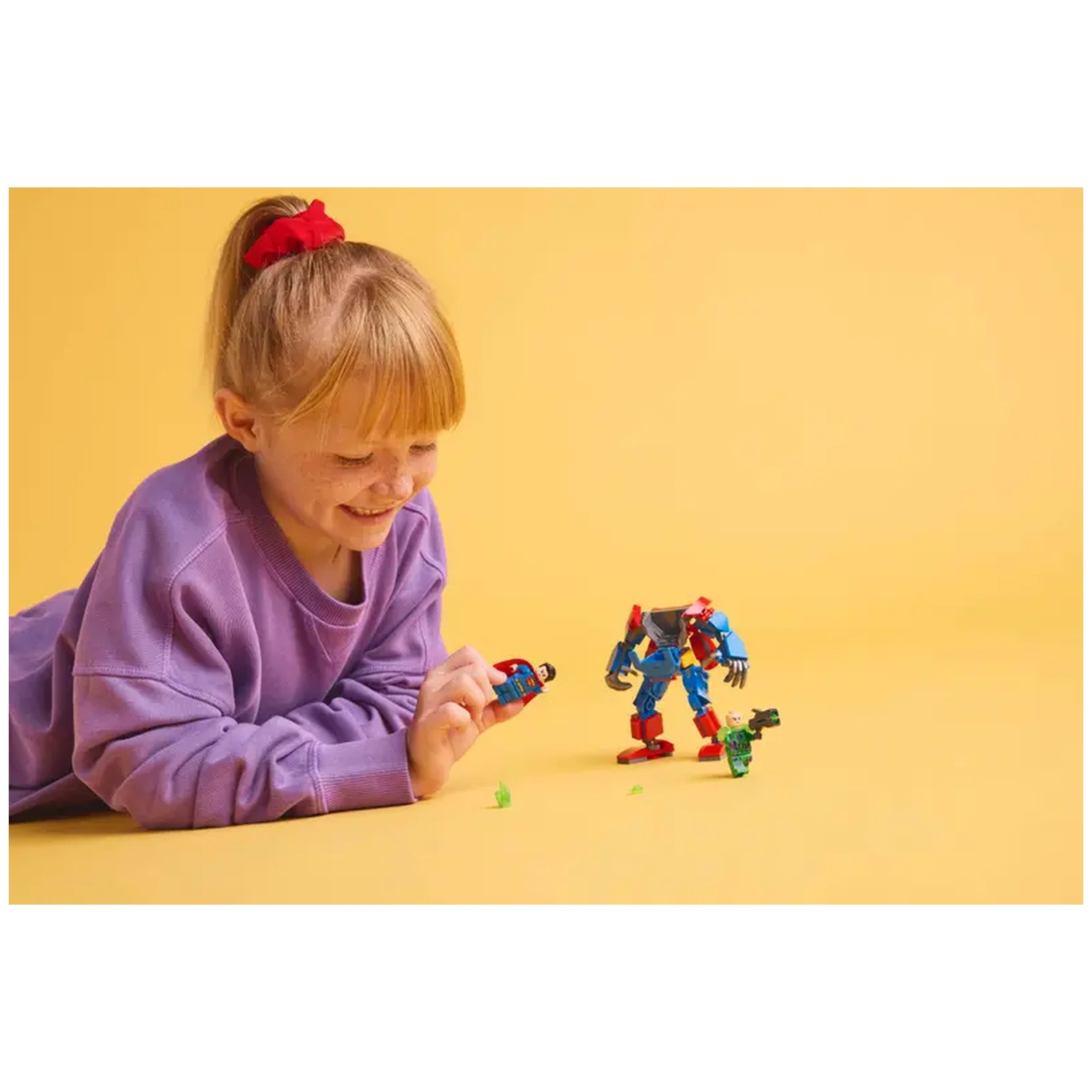 A smiling girl in a purple sweater lays on her stomach, holding a Superman minifigure while playing with a colorful Superman mech and Lex Luthor figure on a bright yellow background.