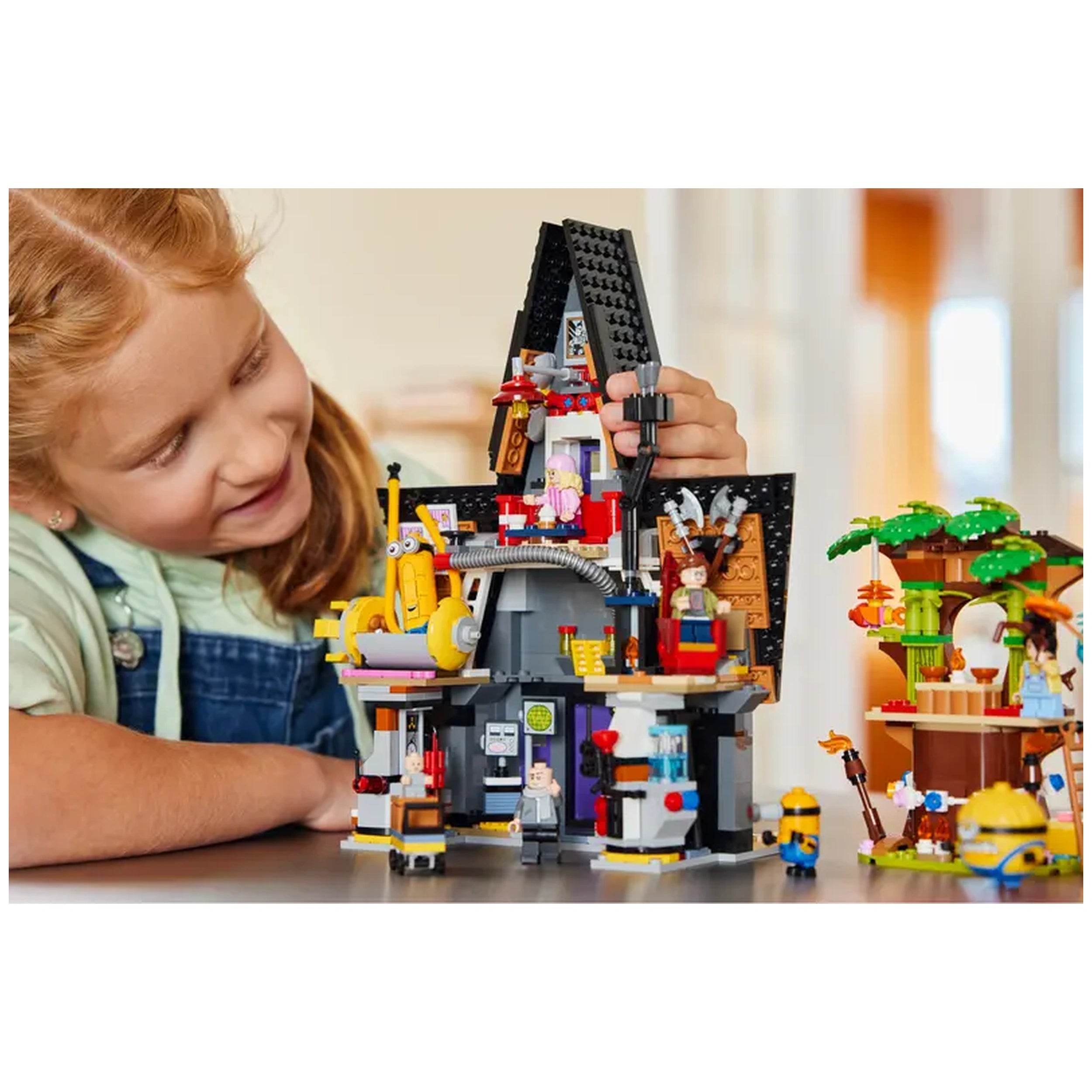 A young girl with curly hair smiles as she plays with the intricate LEGO® Despicable Me construction set, showcasing Gru's mansion and colorful Minions figures. The set features detailed rooms, including a lab and cozy spaces, all vibrantly designed, encouraging imaginative play.