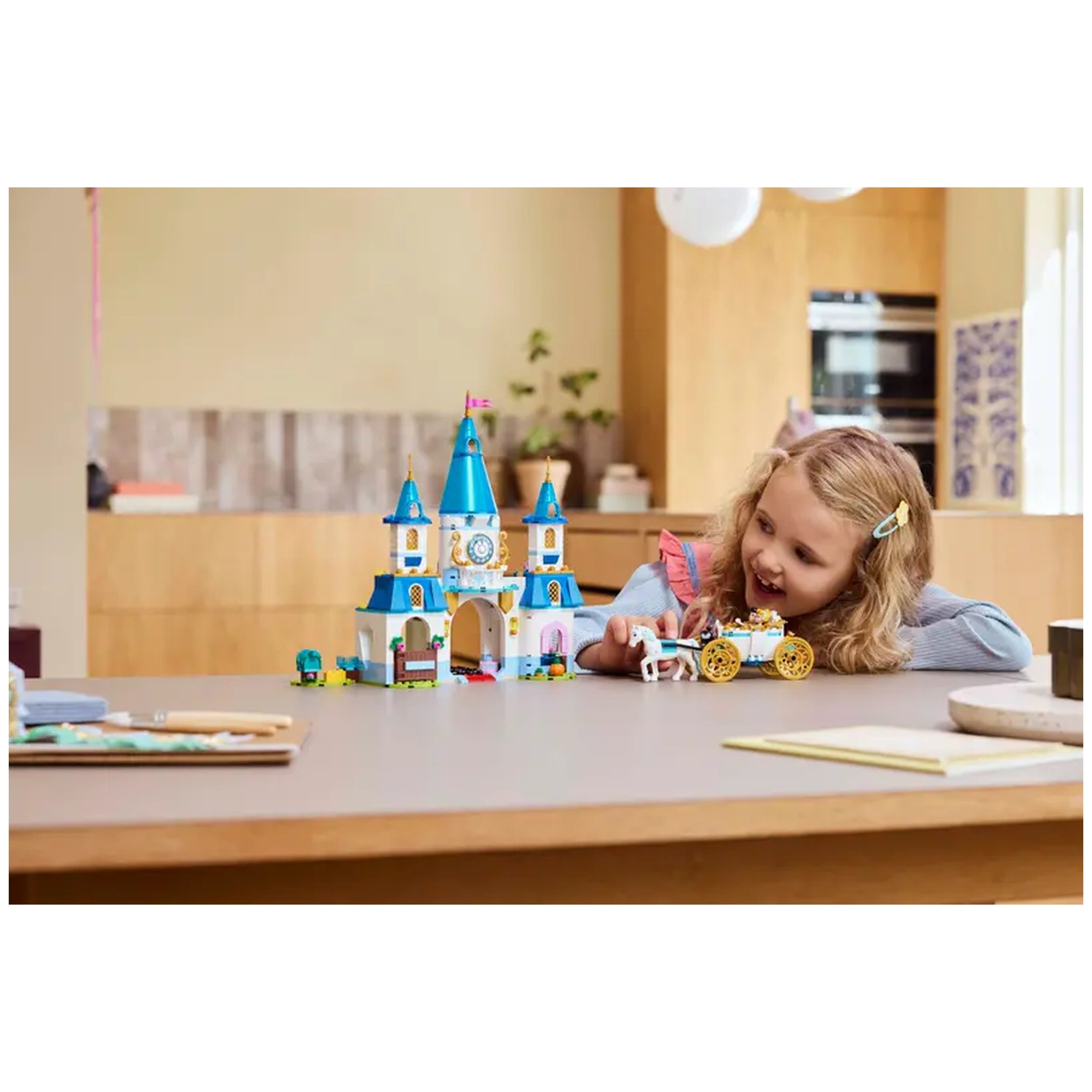 A young girl with curly hair smiles while playing with the LEGO® Disney Princess Cinderella’s Castle set, featuring a blue-roofed castle and a white horse-drawn carriage, on a wooden table in a bright, modern kitchen.