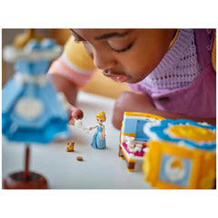 A child focuses intently on a LEGO® Cinderella minidoll dressed in a blue gown, serving tea to a small brown mouse beside a colorful building set, with a blue lamp in the background.