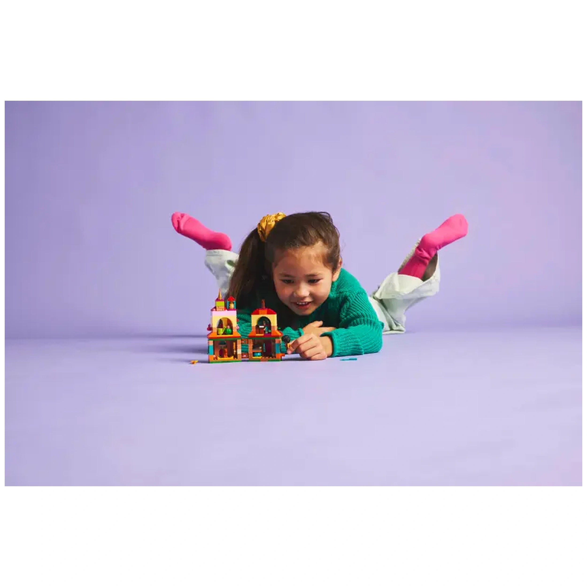 A young girl wearing a green sweater and pink socks is lying on her stomach, focused on playing with the LEGO® Disney Encanto Mini House set on a light purple background.