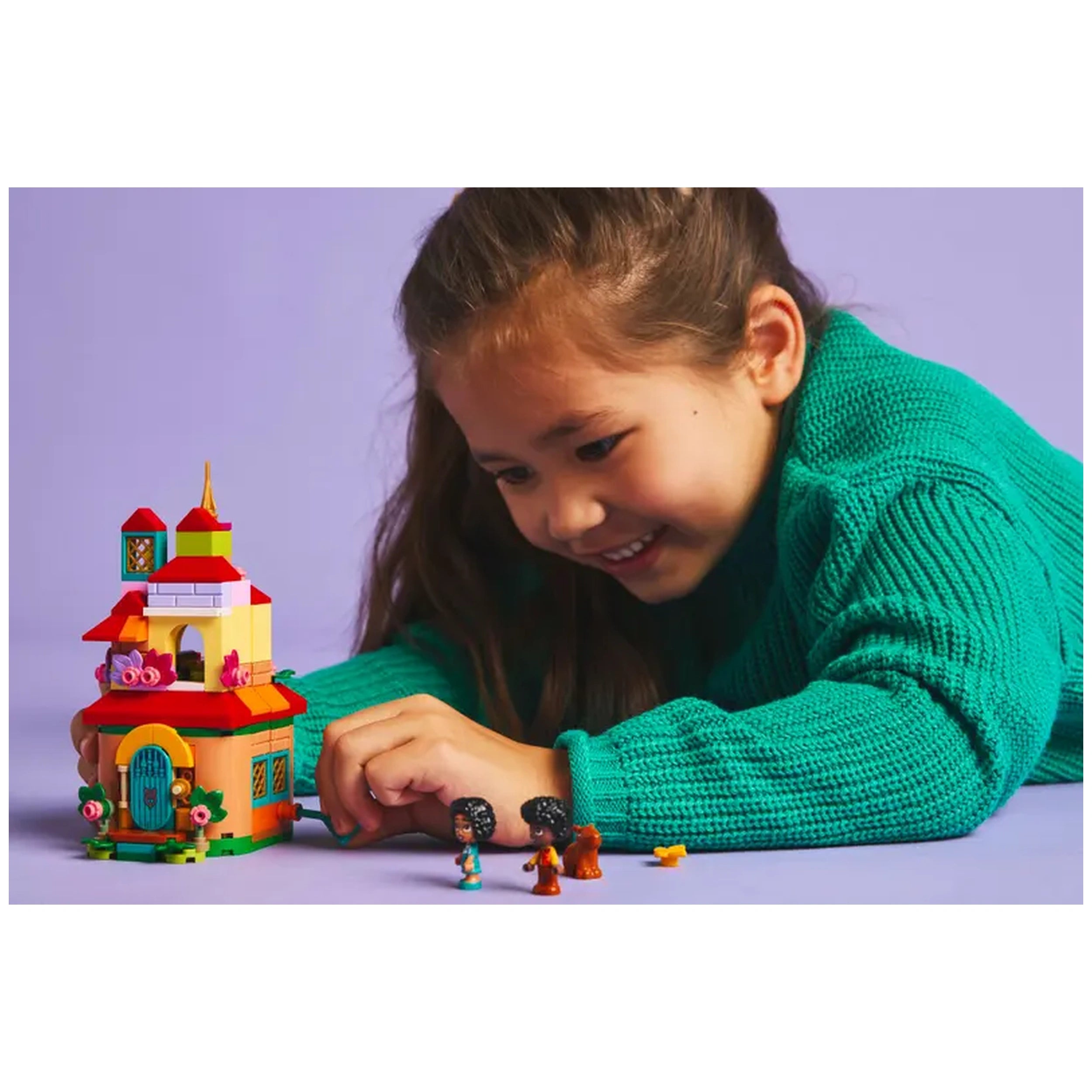 A child in a green sweater smiles while playing with the LEGO Disney Encanto Mini House set, featuring vibrant colors and floral details. Next to the house, LEGO characters including Mirabel and Antonio are arranged for imaginative play.
