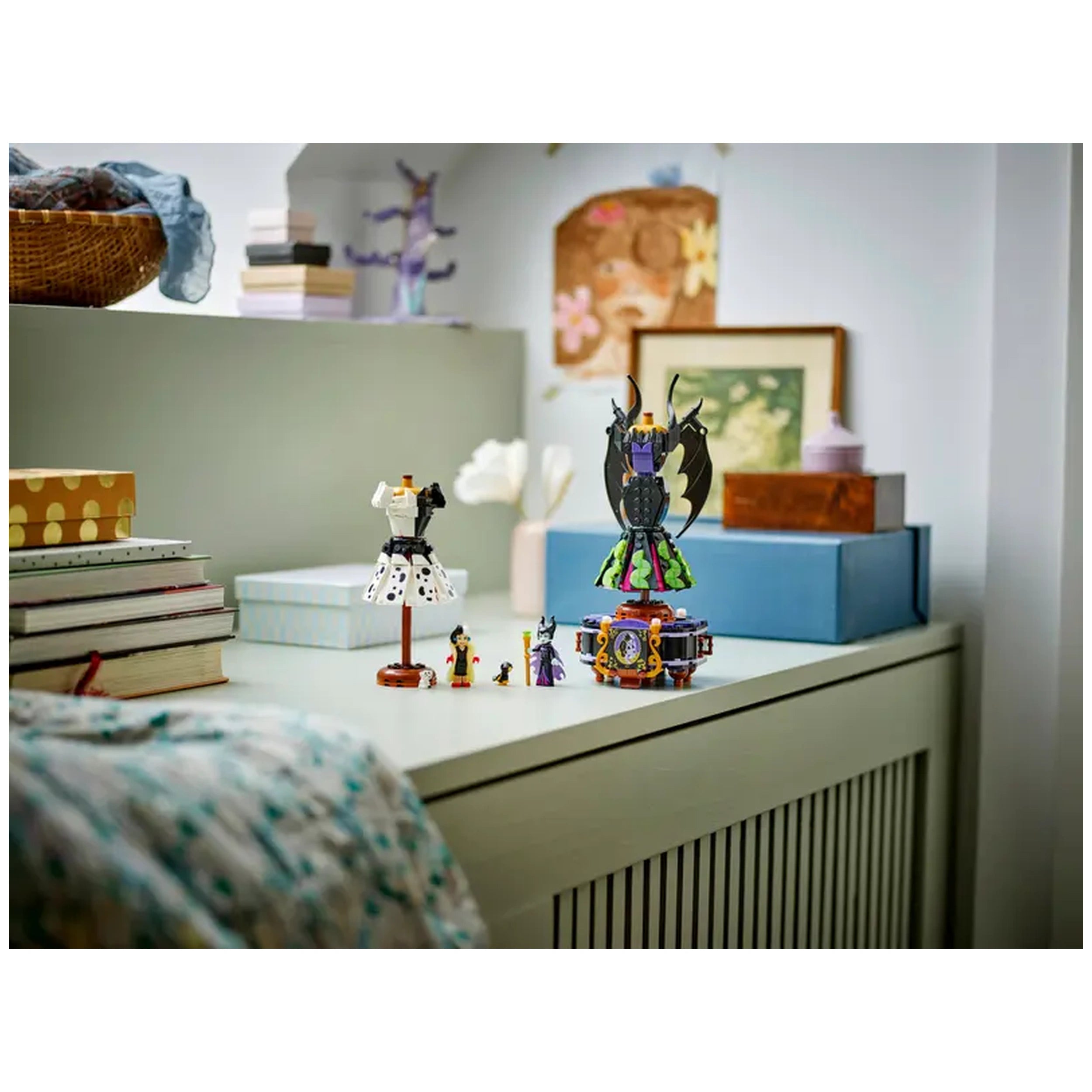 Two buildable dresses representing Maleficent and Cruella De Vil displayed on a dresser, surrounded by LEGO minidolls and accessories, with books and decorative items in the background.