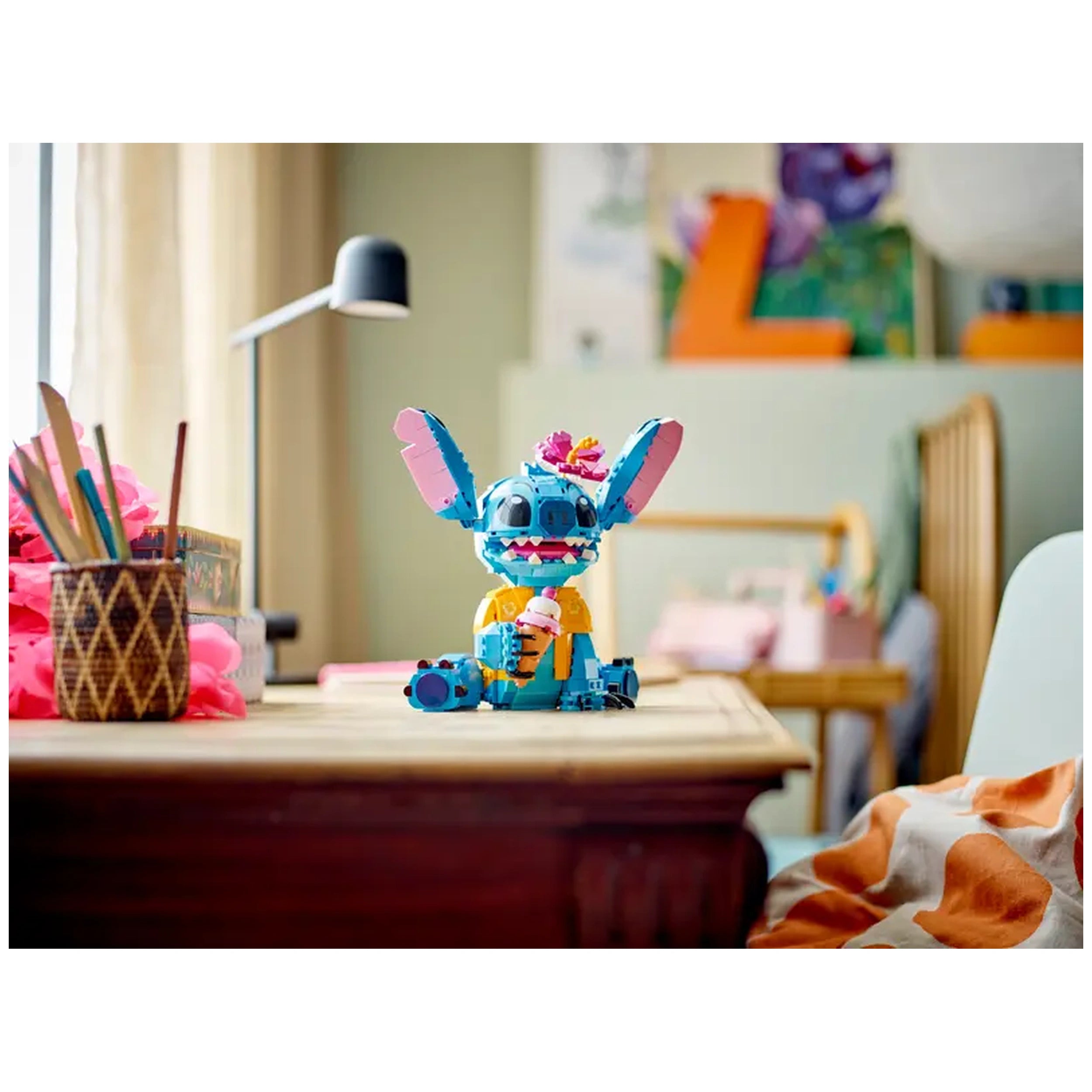 LEGO® Disney Stitch construction set on a wooden table, vibrant blue Stitch figure with movable ears, holding an ice cream cone, surrounded by colorful desk accessories and a cozy room background.