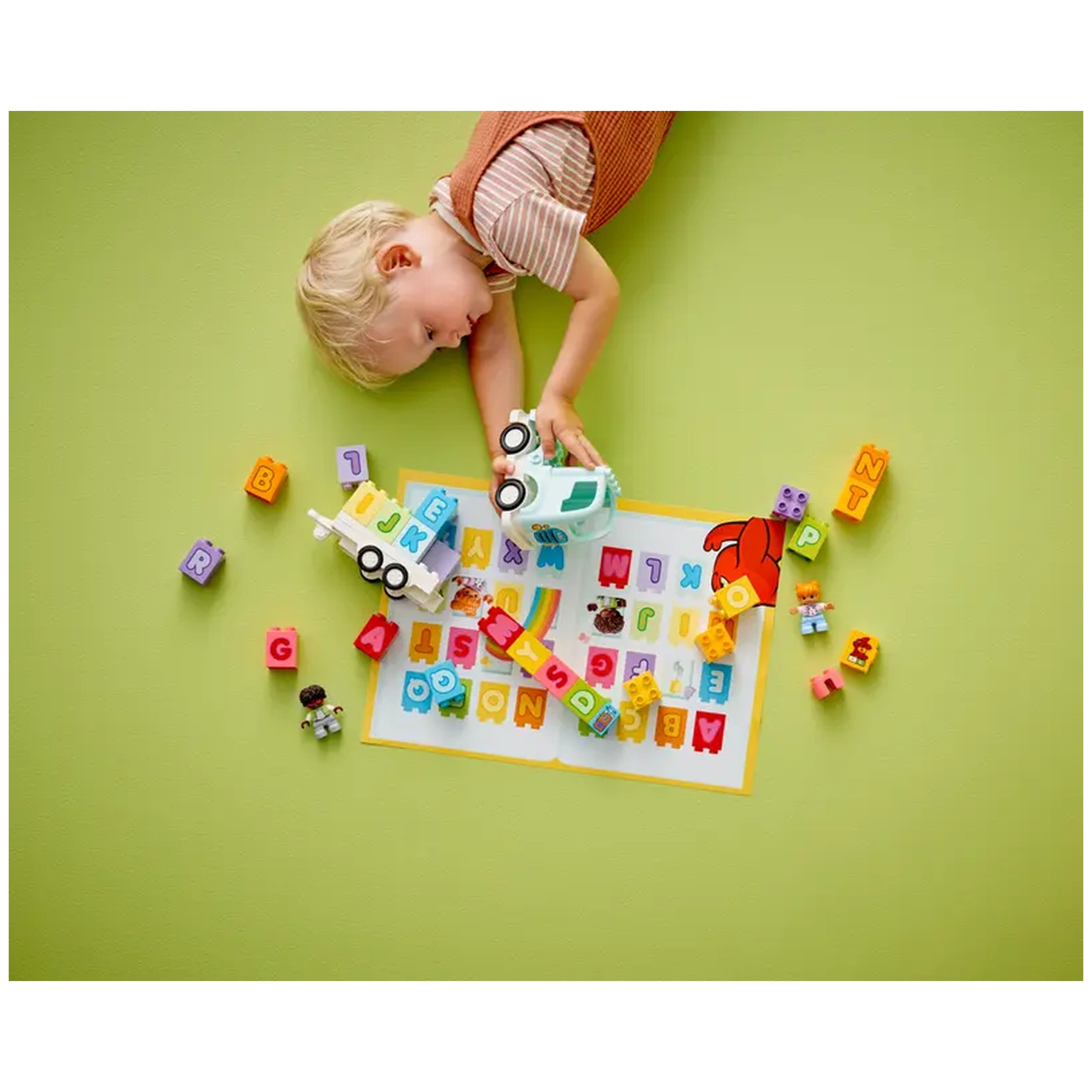 A toddler plays on a green floor, interacting with a white truck toy and colorful alphabet blocks scattered around.