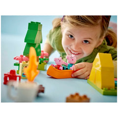 Young girl smiling while playing with the LEGO® DUPLO® Camping Trip set, featuring Peppa Pig and Daddy Pig in a canoe surrounded by colorful blocks and flowers.