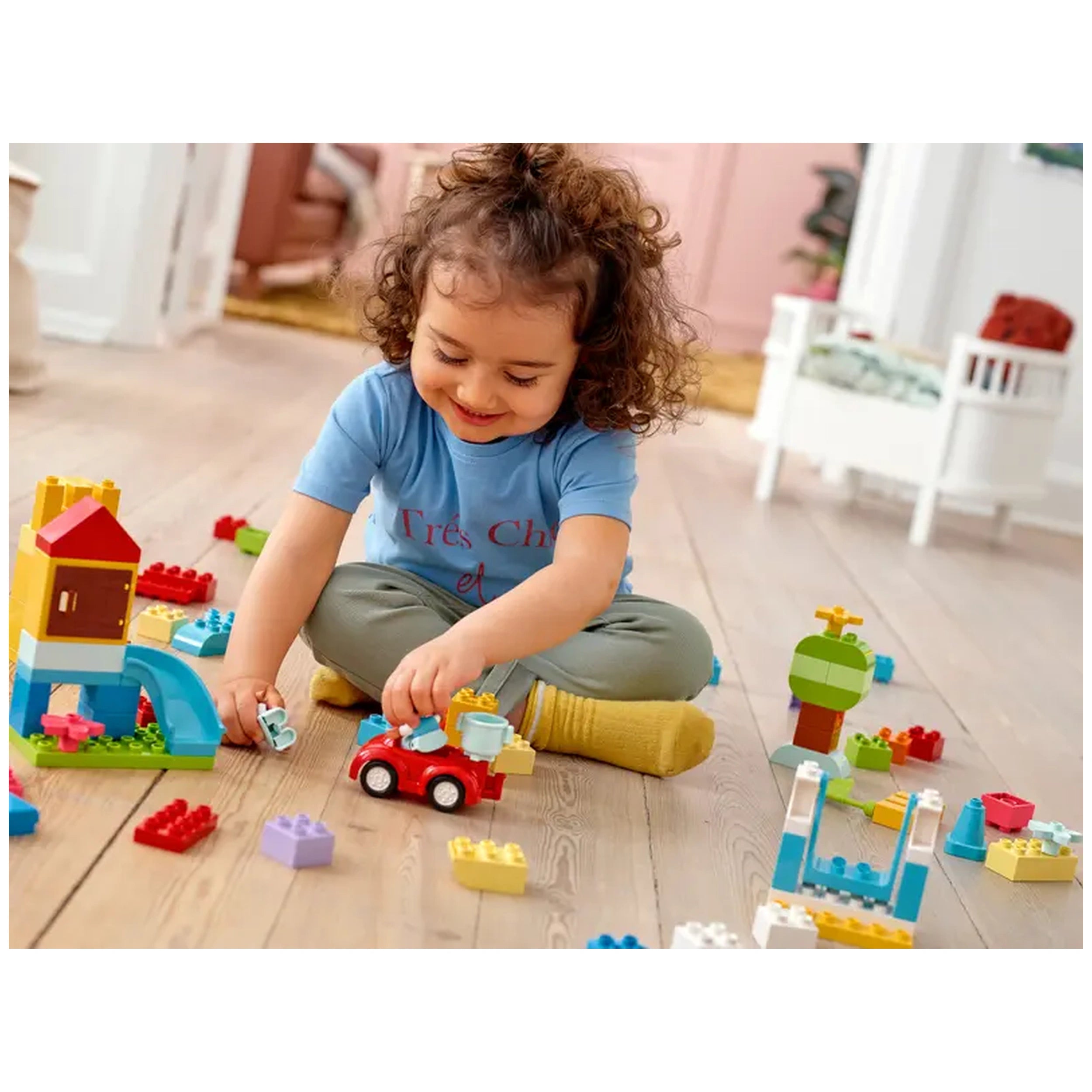 A joyful toddler with curly hair sits on the floor, playing with colorful LEGO® DUPLO® bricks. The child is assembling a red toy car, surrounded by various building pieces, including a small house and a tree. Bright, cheerful colors enhance the playful atmosphere in a cozy room adorned with soft furnishings.