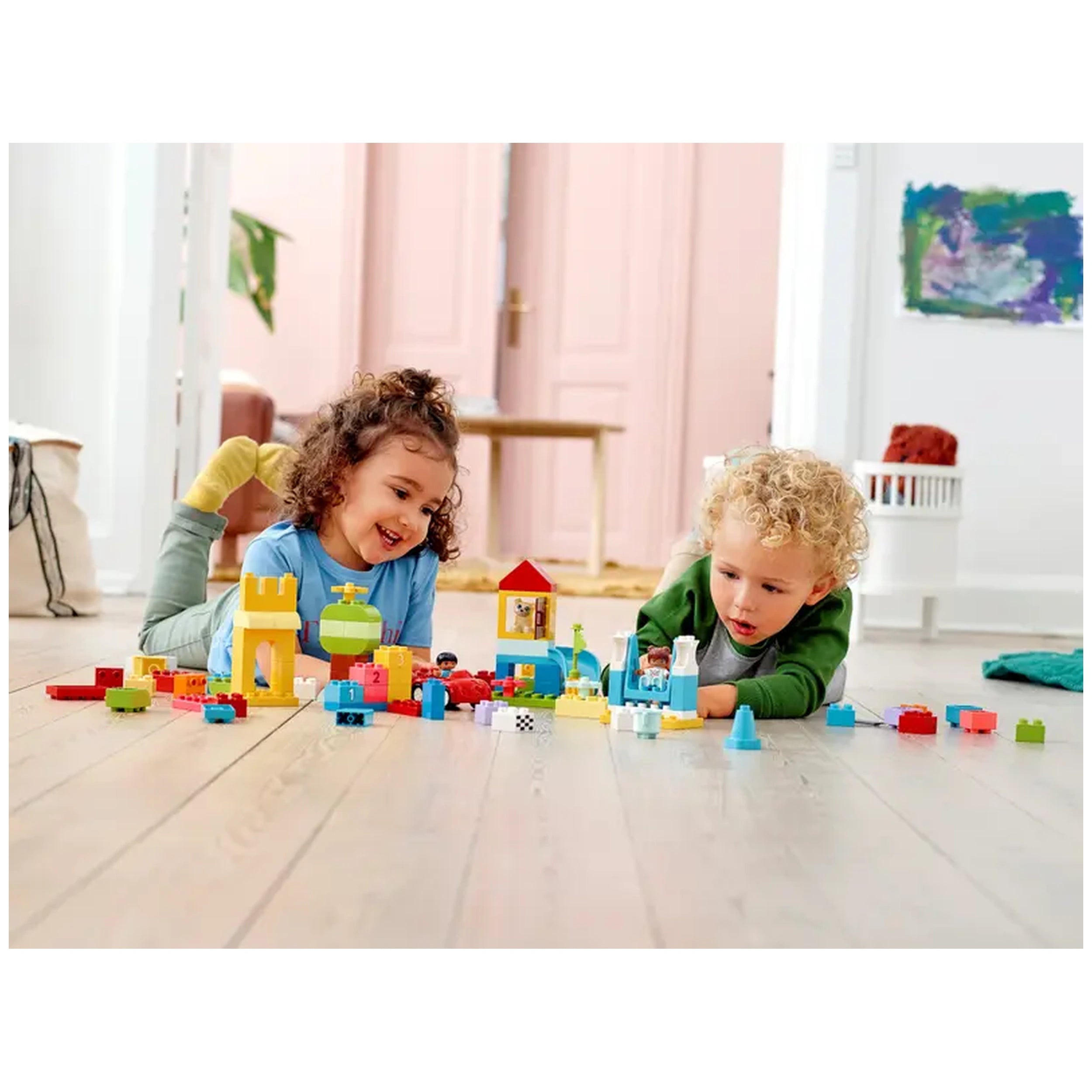 Two young children play on a wooden floor with colorful LEGO® DUPLO® bricks, building a castle and other structures. The girl smiles, wearing a blue shirt, while the boy, in a green sweater, focuses intently on his creation. There are vibrant pieces scattered around, encouraging imaginative play in a bright, cheerful room.