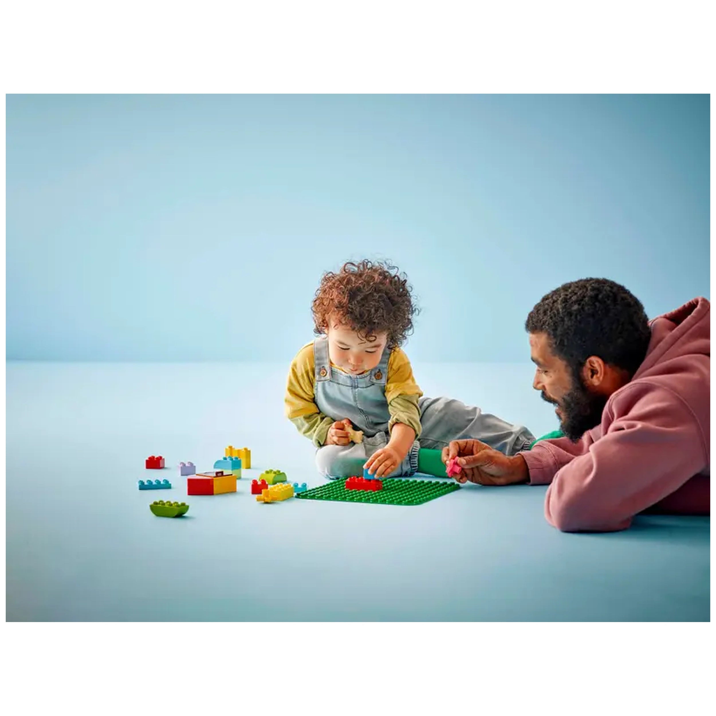 A preschool child and a man play together on a light blue surface, building with colorful LEGO® DUPLO® bricks on a green baseplate. The child, wearing a yellow shirt and denim overalls, focuses on placing a red piece, while the man, in a pink hoodie, gently guides him with a pink brick in hand.