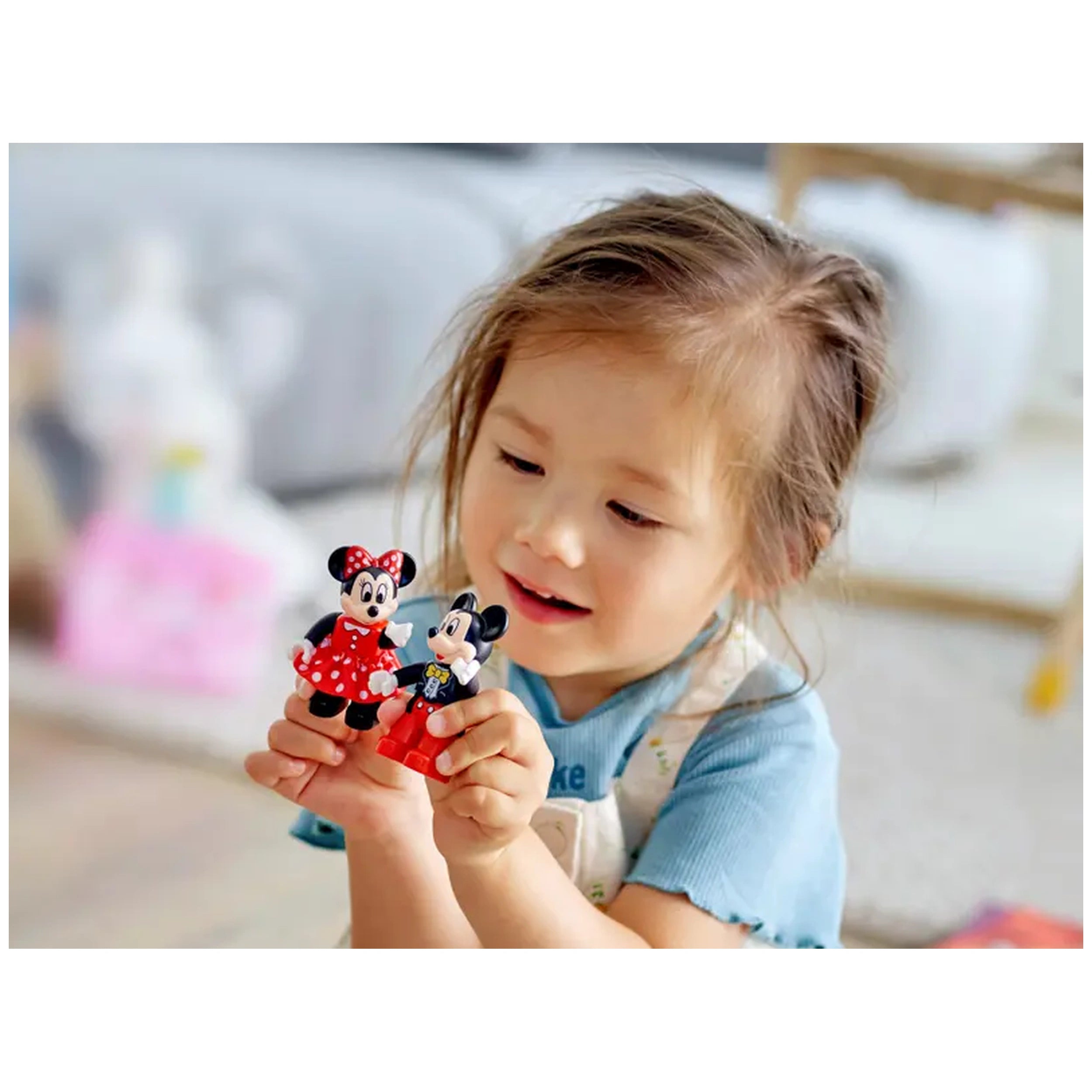 A smiling child holds LEGO® DUPLO® figures of Mickey and Minnie Mouse, dressed in their iconic outfits, in a bright playroom.