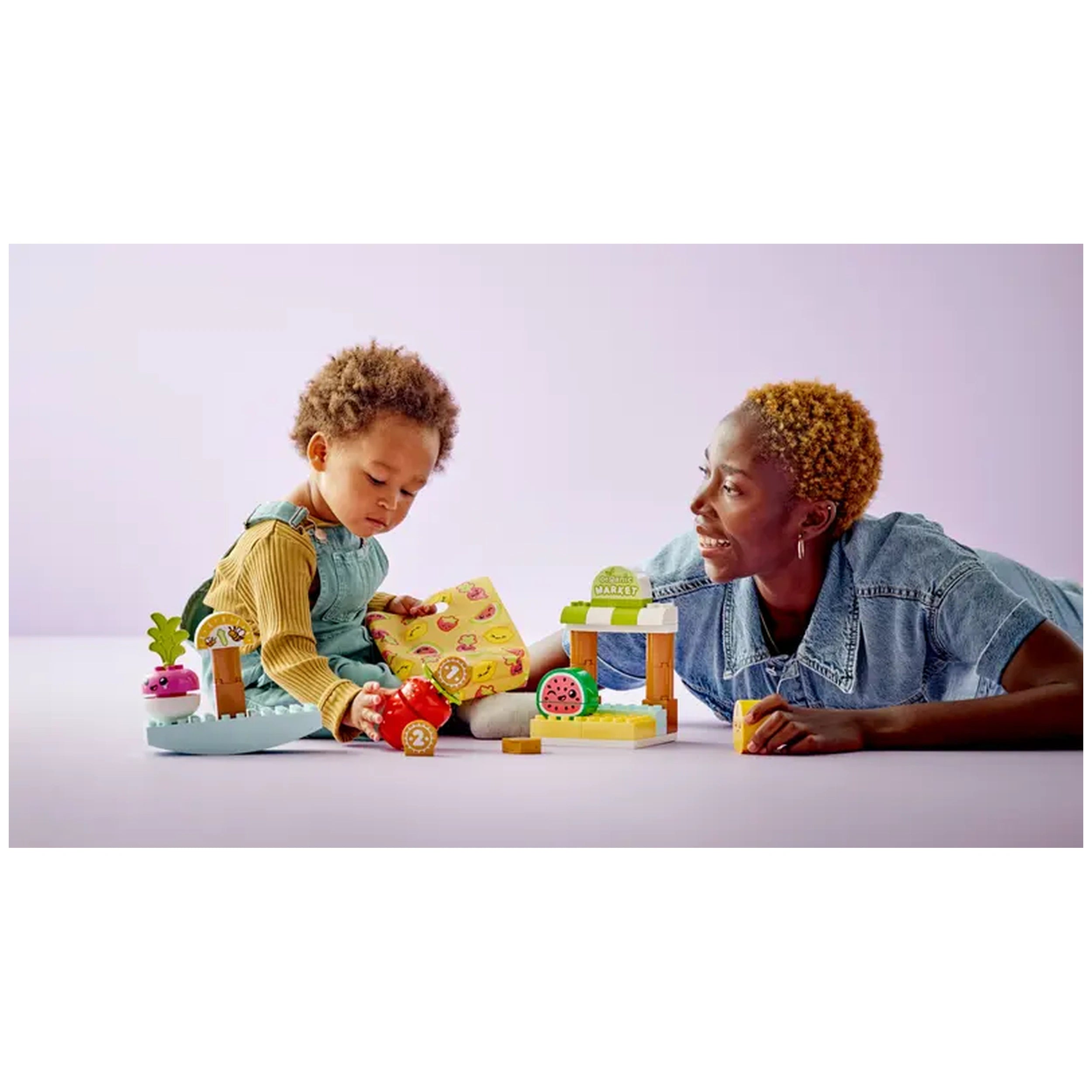Toddlers play with the LEGO® DUPLO® My First Organic Market set, featuring colorful fruits and coins, while an adult engages joyfully with them.
