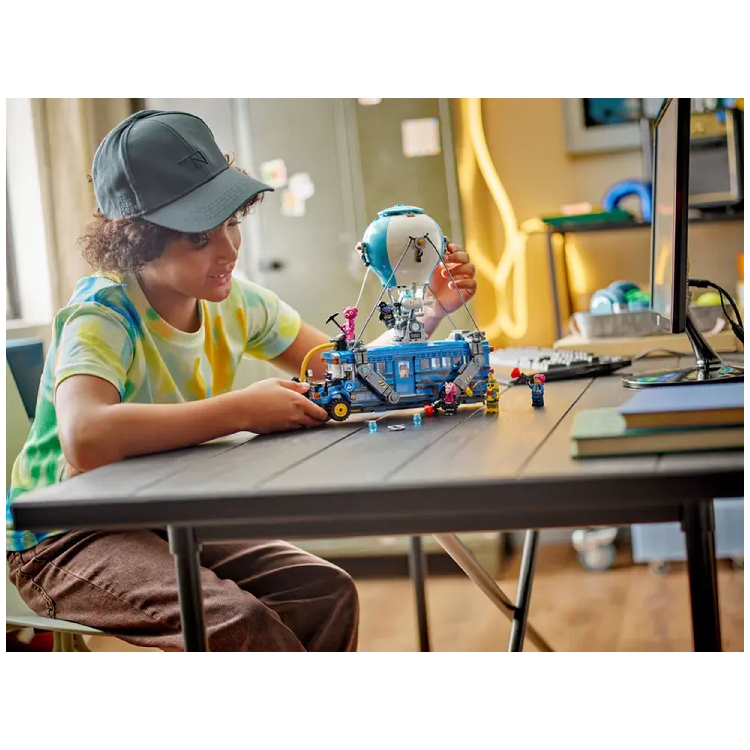 A child wearing a colorful tie-dye shirt and a cap joyfully interacts with a LEGO Fortnite Battle Bus set on a wooden table, surrounded by mini figures and a hot-air balloon.