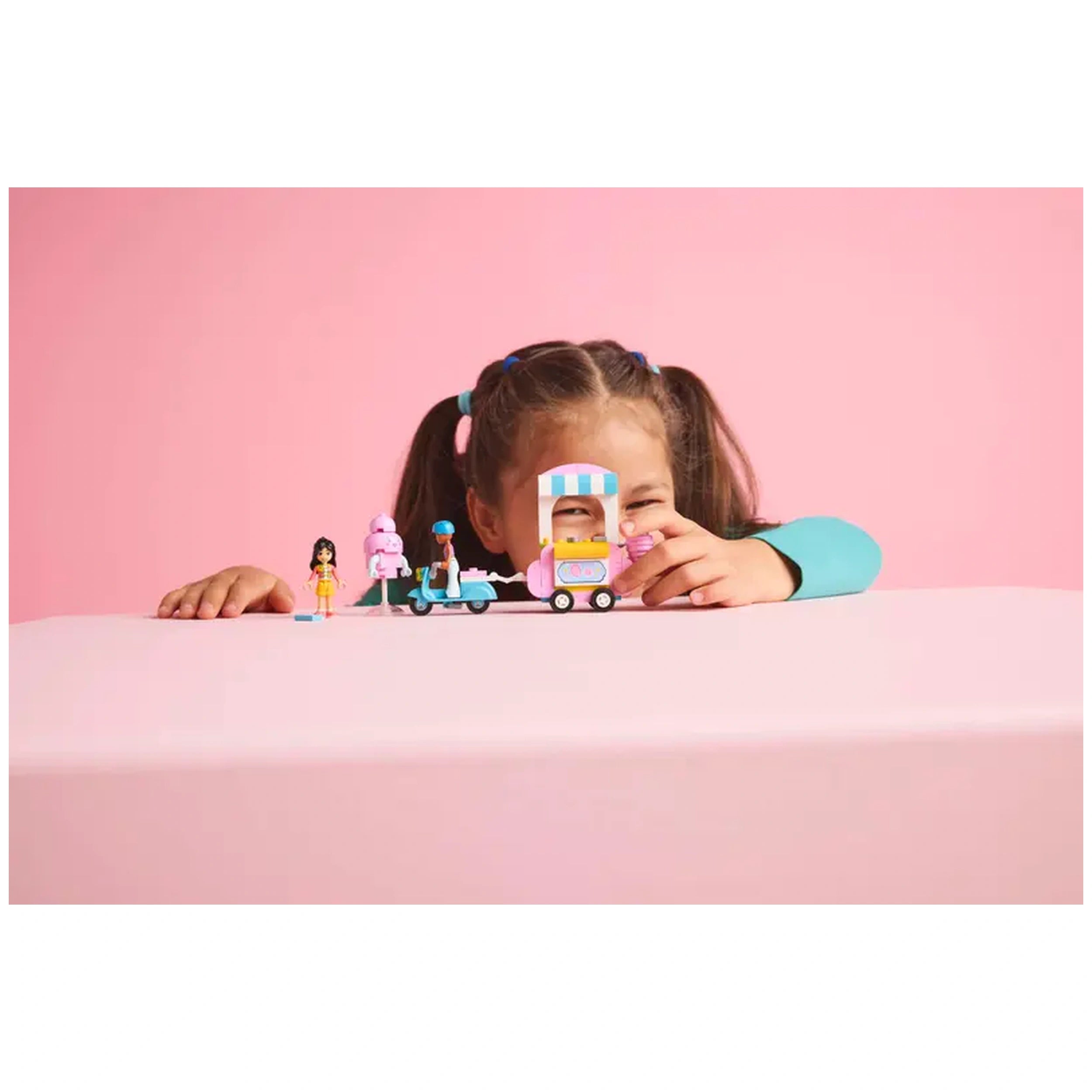 A smiling child with brown hair and pigtails plays with the LEGO® Friends Cotton Candy Stand and Scooter set, featuring two minidolls and a pastel-colored food cart against a pink background.
